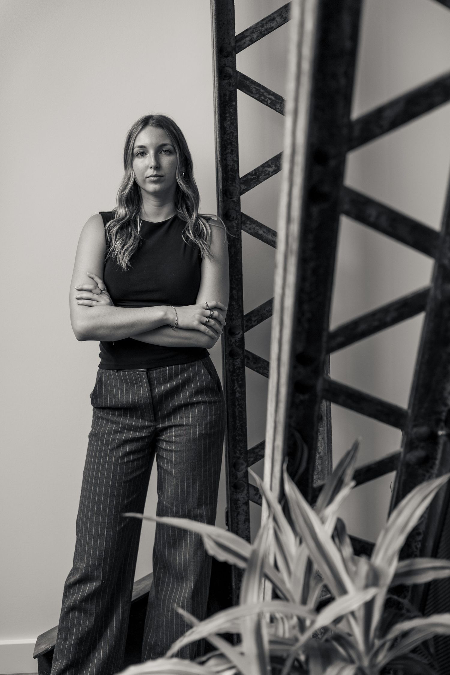 Black and white portrait of a confident woman in striped trousers leaning against a metal structure.