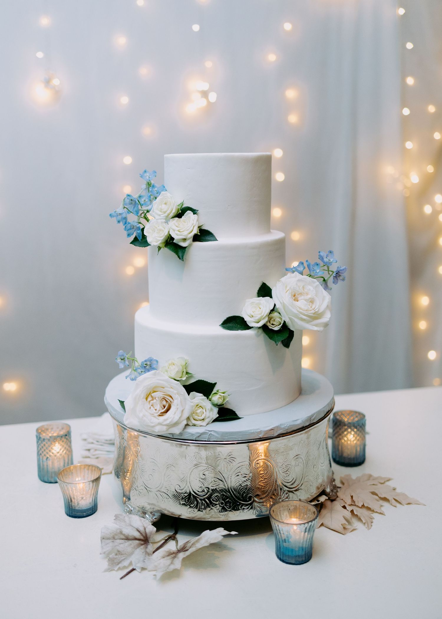 Three-tier white wedding cake with white roses and blue flowers on a silver stand, surrounded by candles and twinkling string lights.