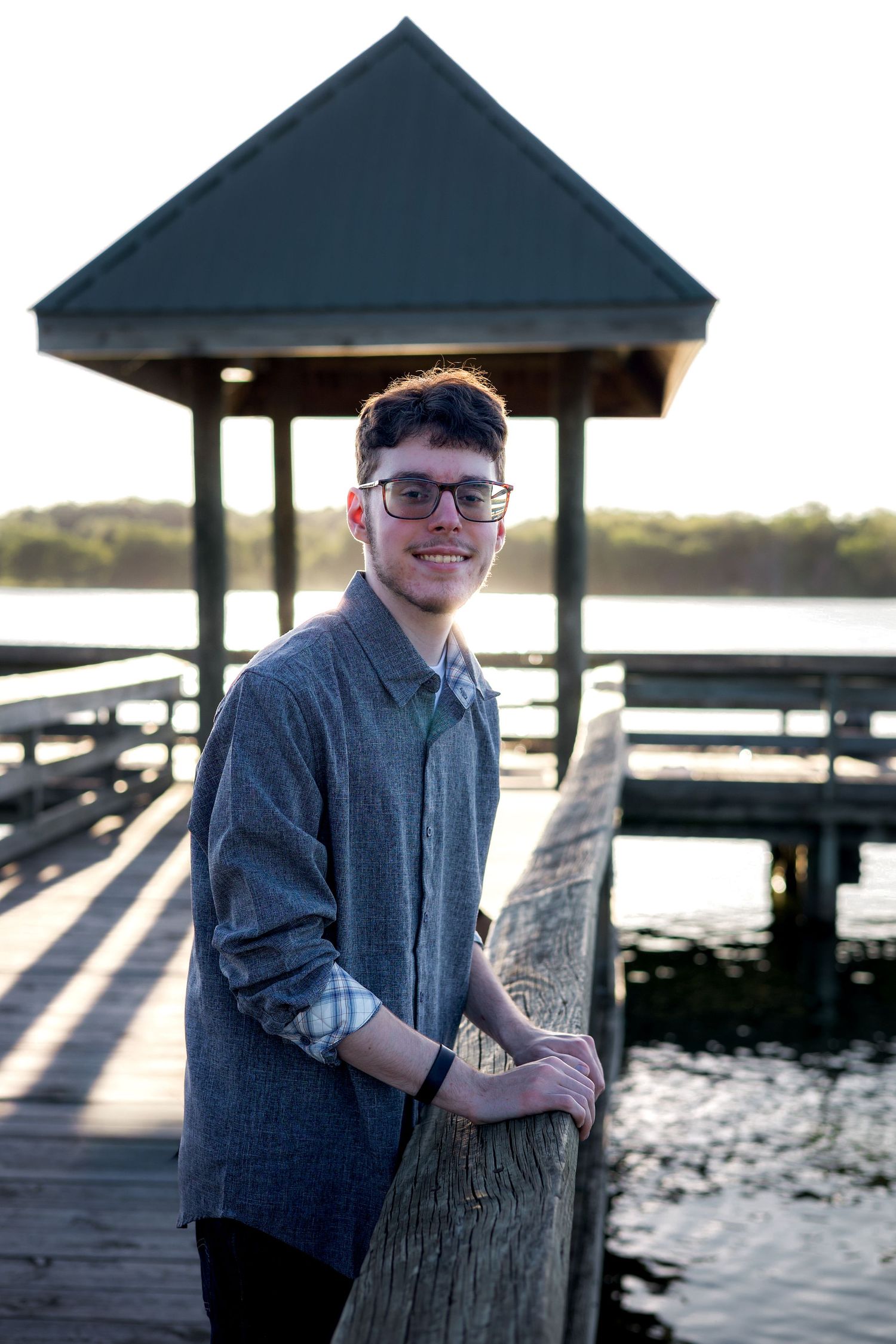 Young man in glasses and gray shirt smiling on a wooden dock by the water at sunset.