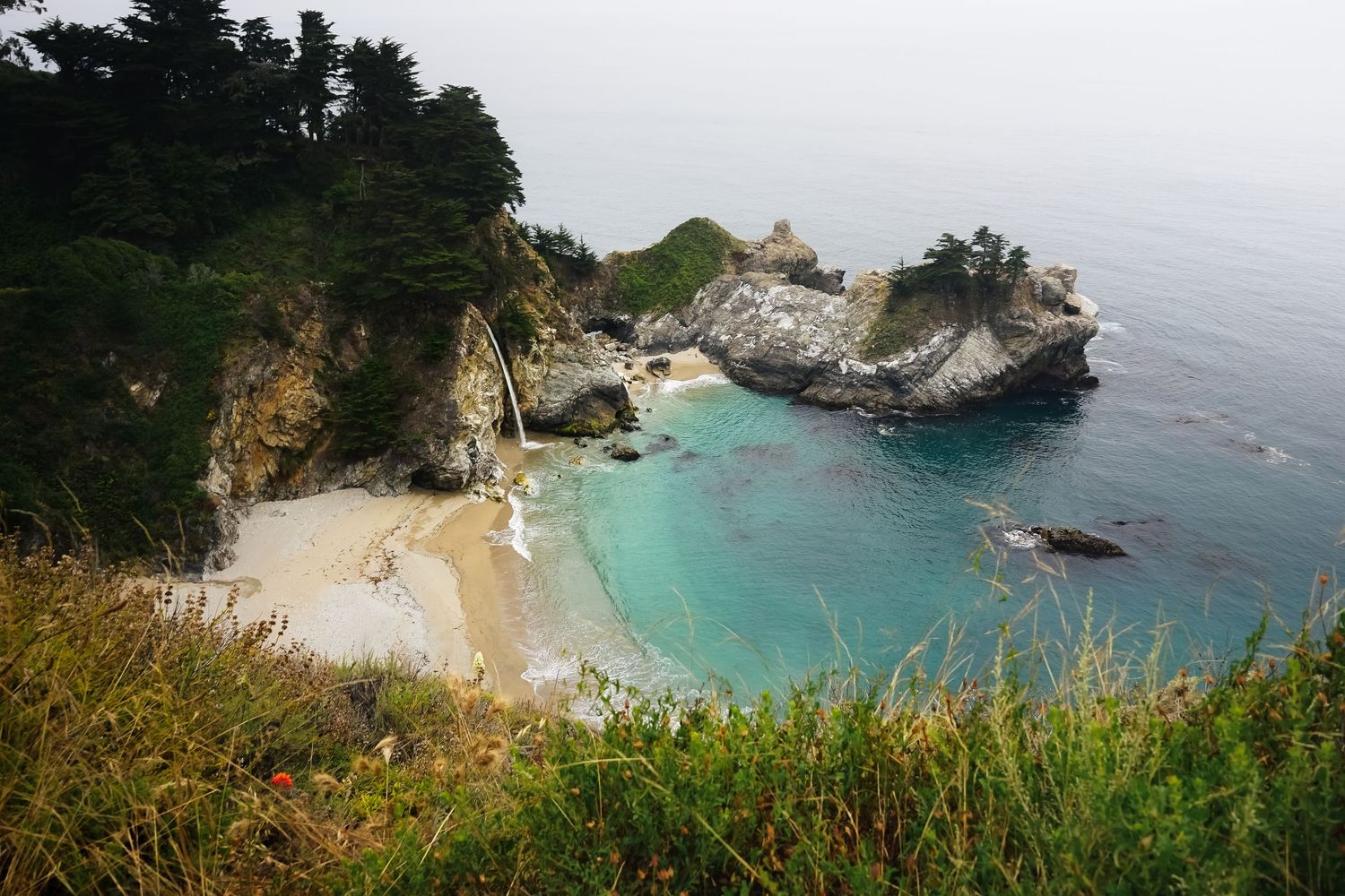 Aerial view of a secluded cove with turquoise water, sandy beach, waterfall, and rocky cliffs at Big Sur, California.