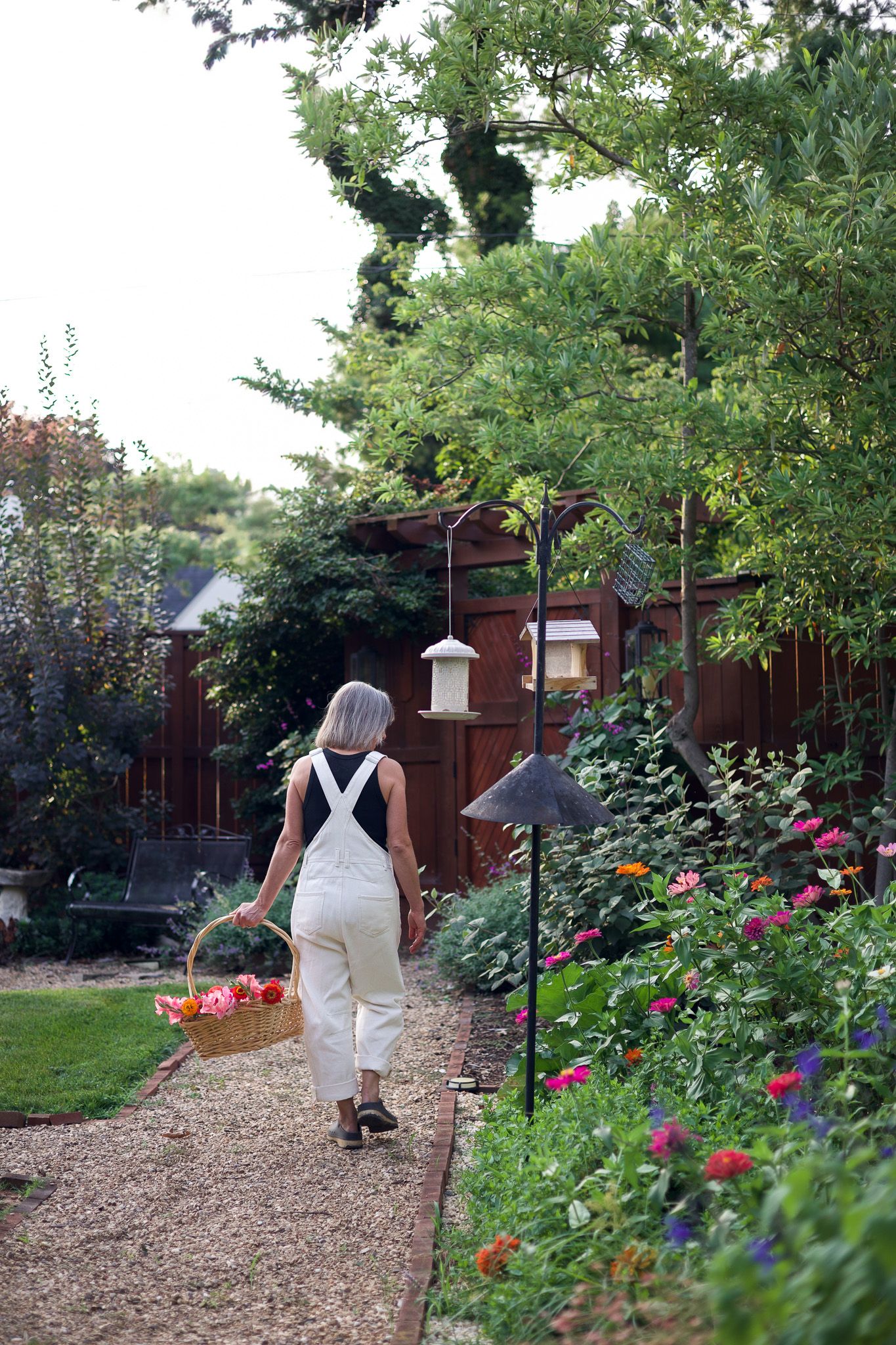 Woman in white overalls carrying a harvest basket along a lush garden path lined with colorful flowers.