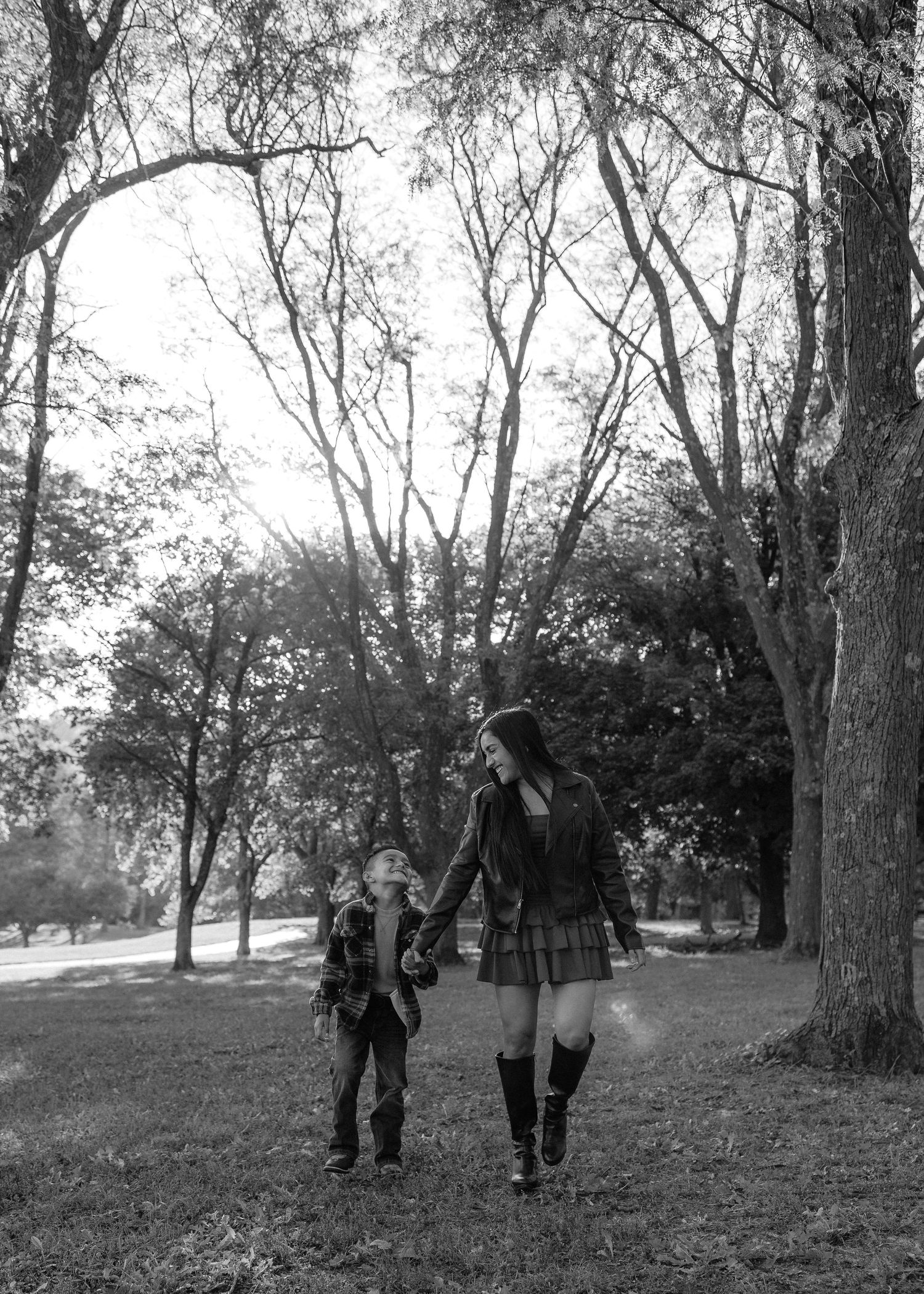 A woman and a young child hold hands while walking through a tree-filled park in this black and white family portrait.