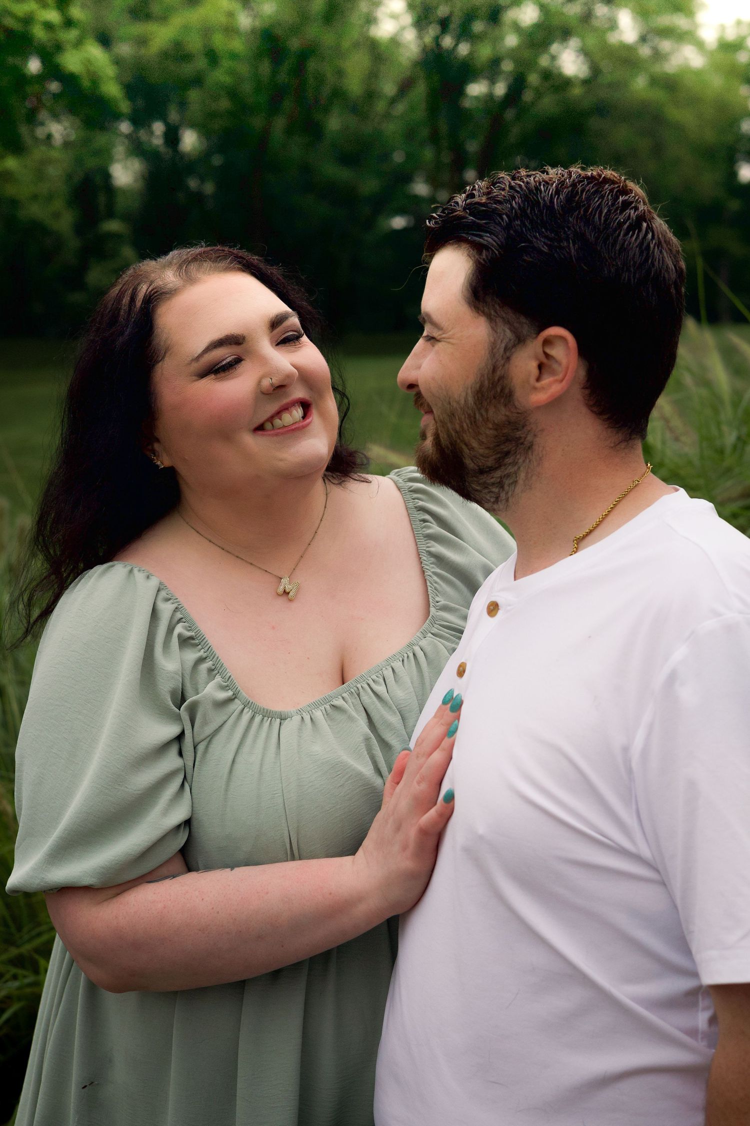 A couple shares a romantic moment outdoors, woman in sage green dress and man in white shirt, surrounded by lush greenery.