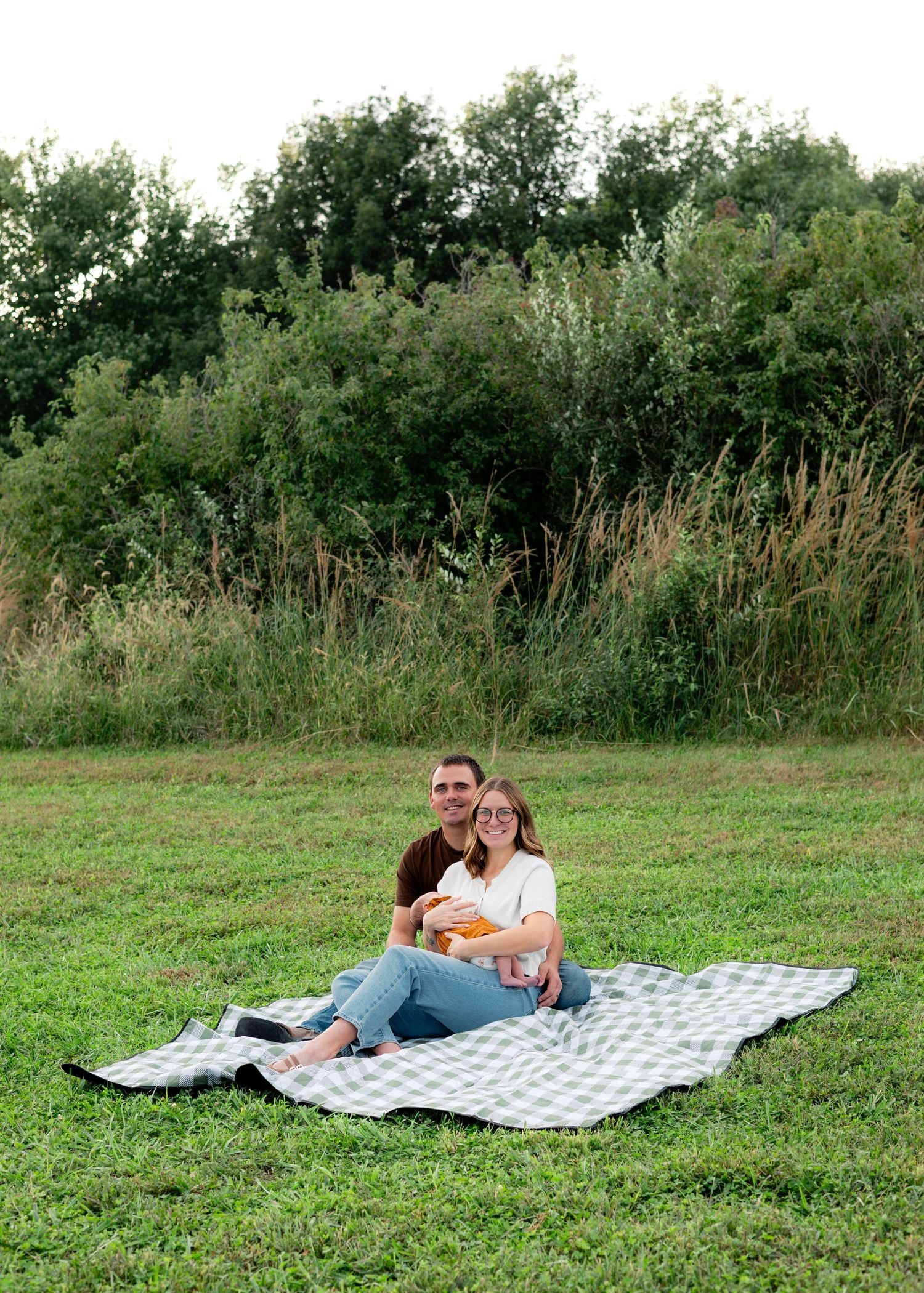Couple sitting on a checkered blanket in a green field, smiling and holding a small pumpkin during an outdoor picnic.