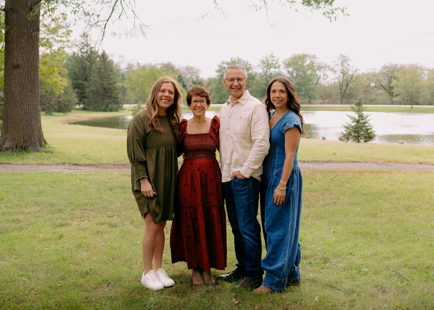 Four adults in coordinated casual outfits pose together on a grassy lawn beside a calm pond with trees in the background.