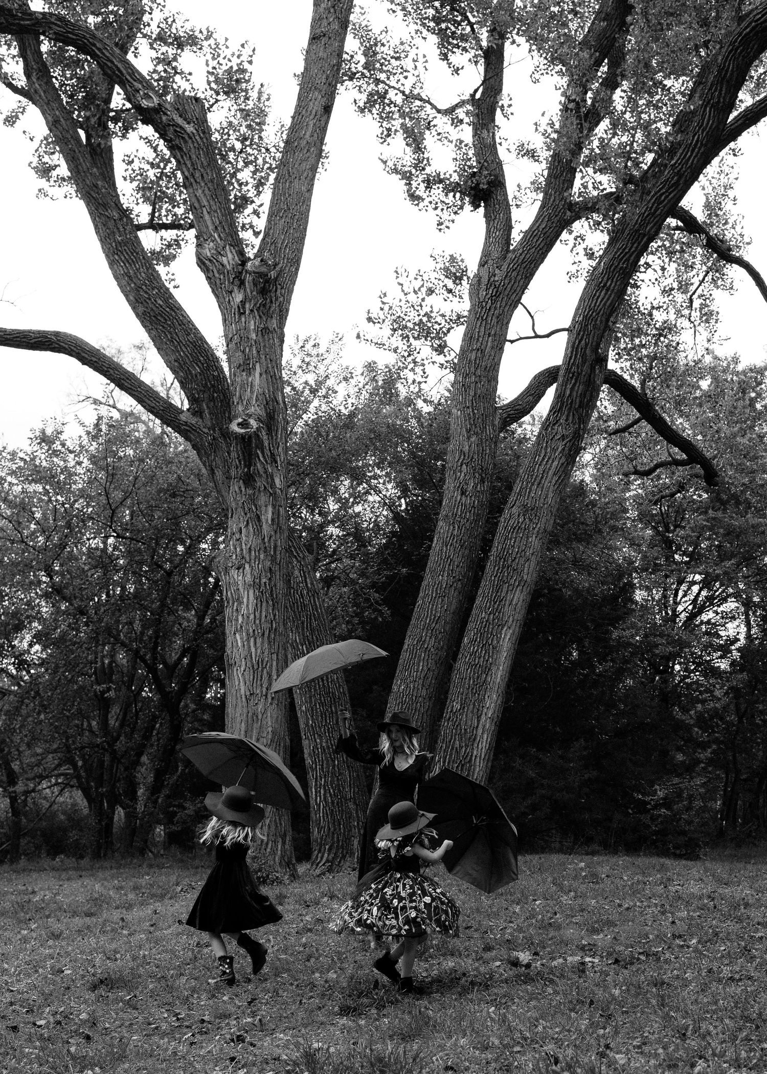 Black and white photo of two children dancing with umbrellas near large trees in a park.
