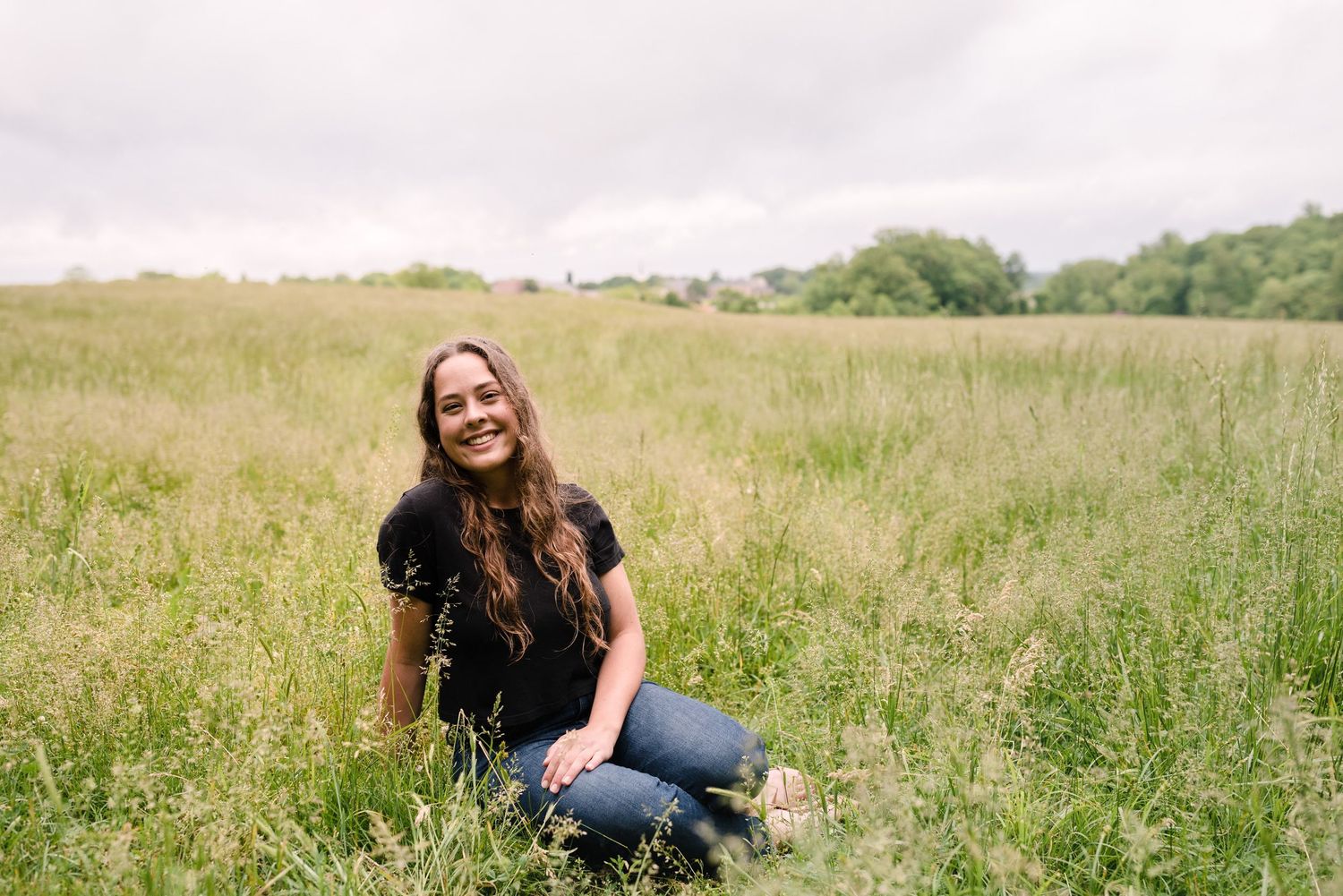 Smiling young woman sitting in a lush green meadow under a cloudy sky.