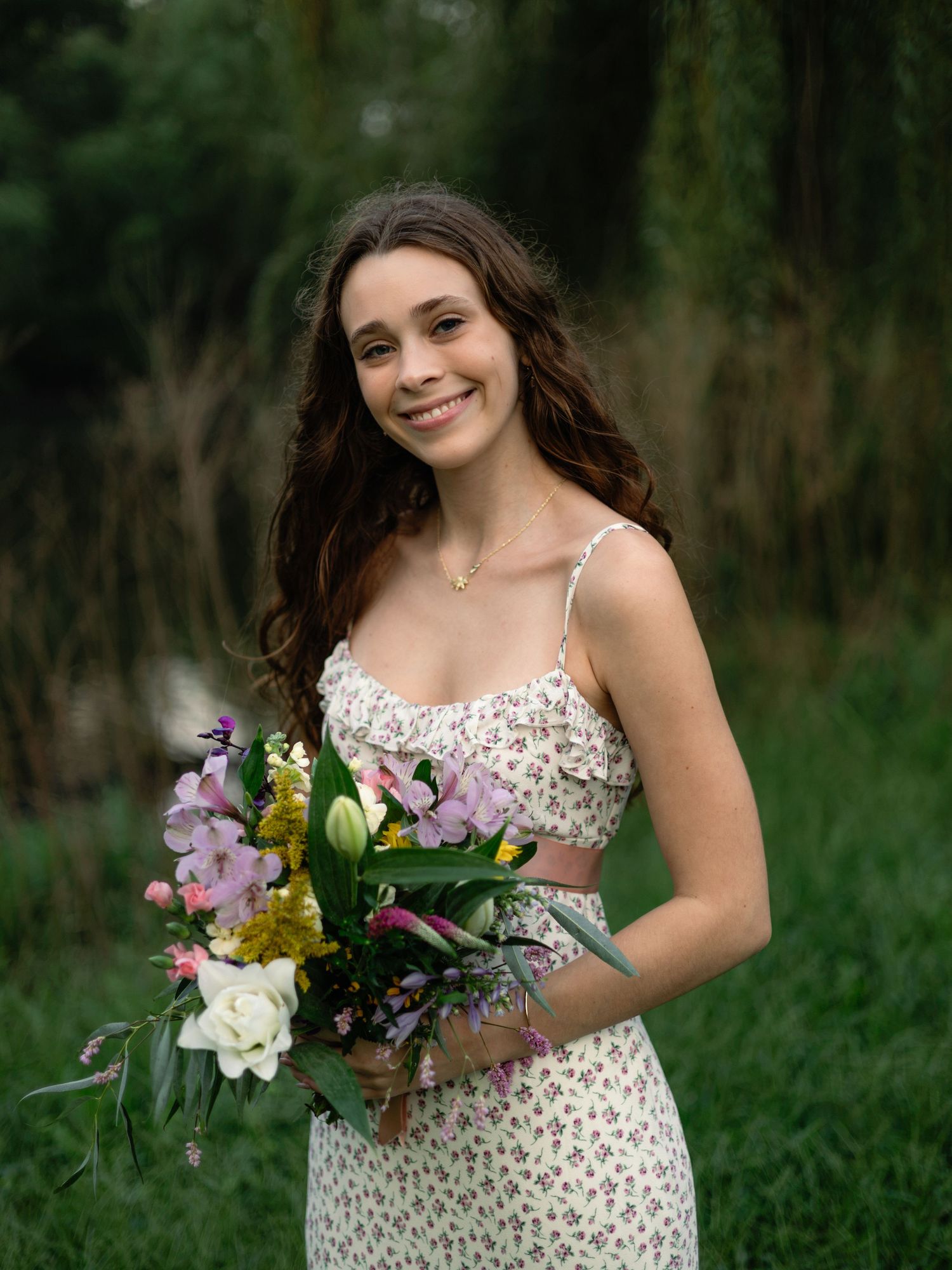 Smiling woman in floral dress holding colorful bouquet of mixed flowers outdoors in lush green garden setting.