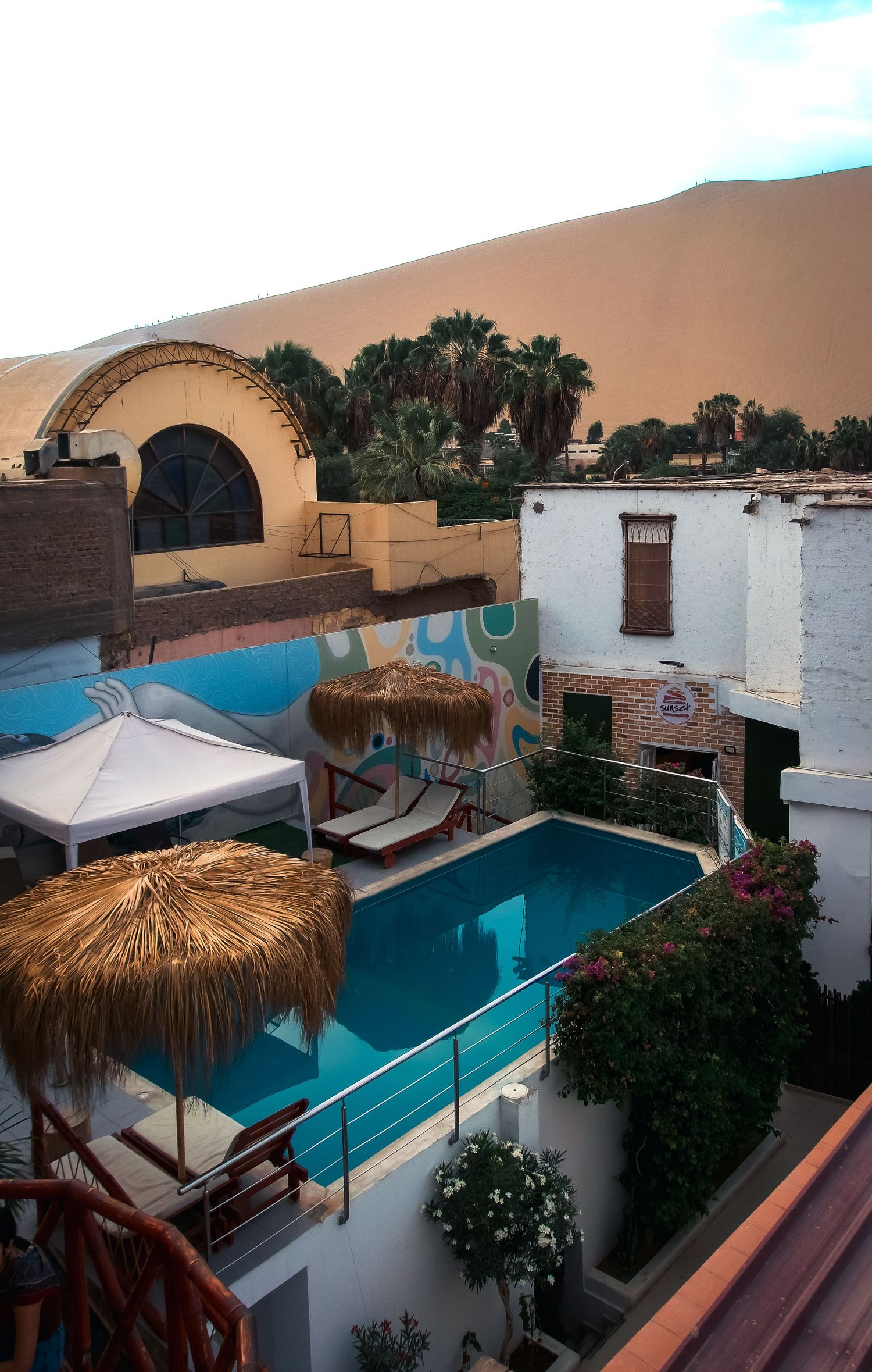 Aerial view of a desert oasis hotel pool with thatched umbrellas, white buildings, palm trees, and massive sand dunes backdrop.