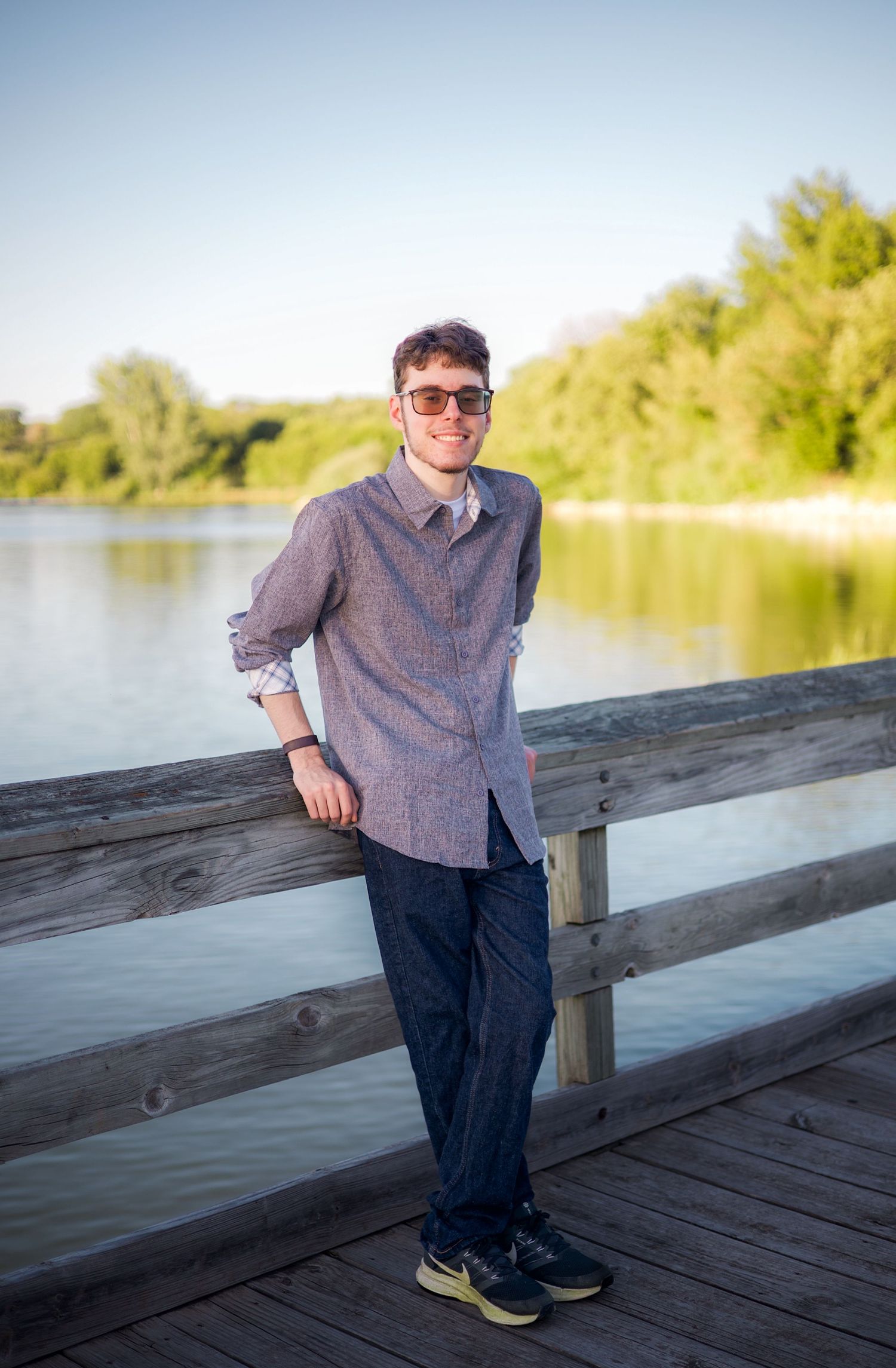 Young man in sunglasses and blue shirt smiling on a wooden dock by a serene lake surrounded by green trees.