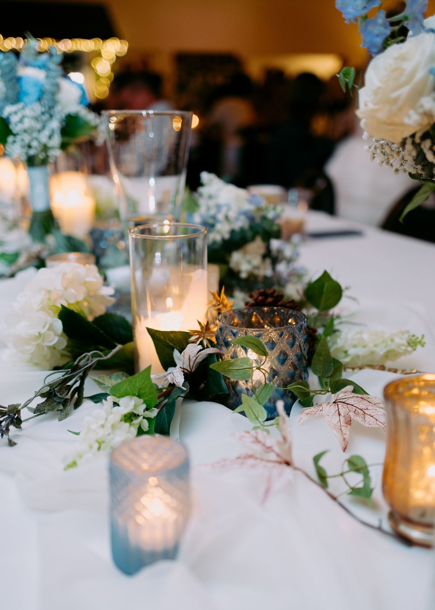 Wedding reception table with flickering votive candles, white and blue floral arrangements, and trailing greenery on a white linen.