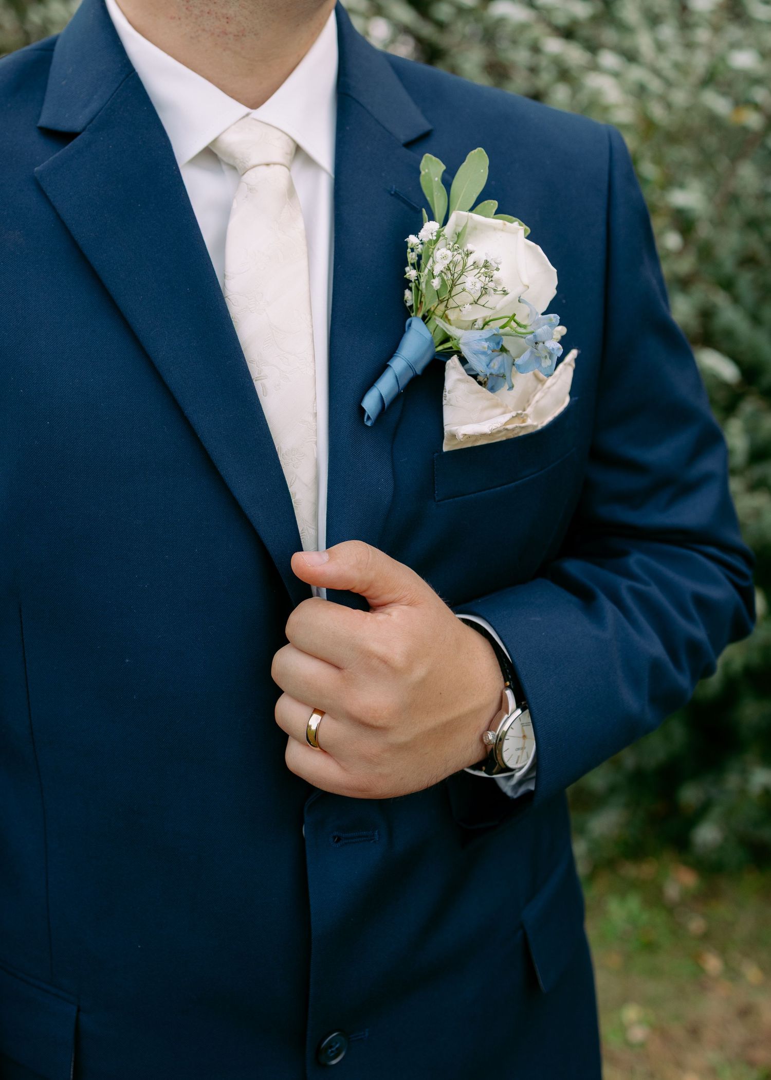 Groom in a navy suit and ivory tie adjusts his jacket, showing a white rose and blue floral boutonniere, wedding band, and wristwatch.