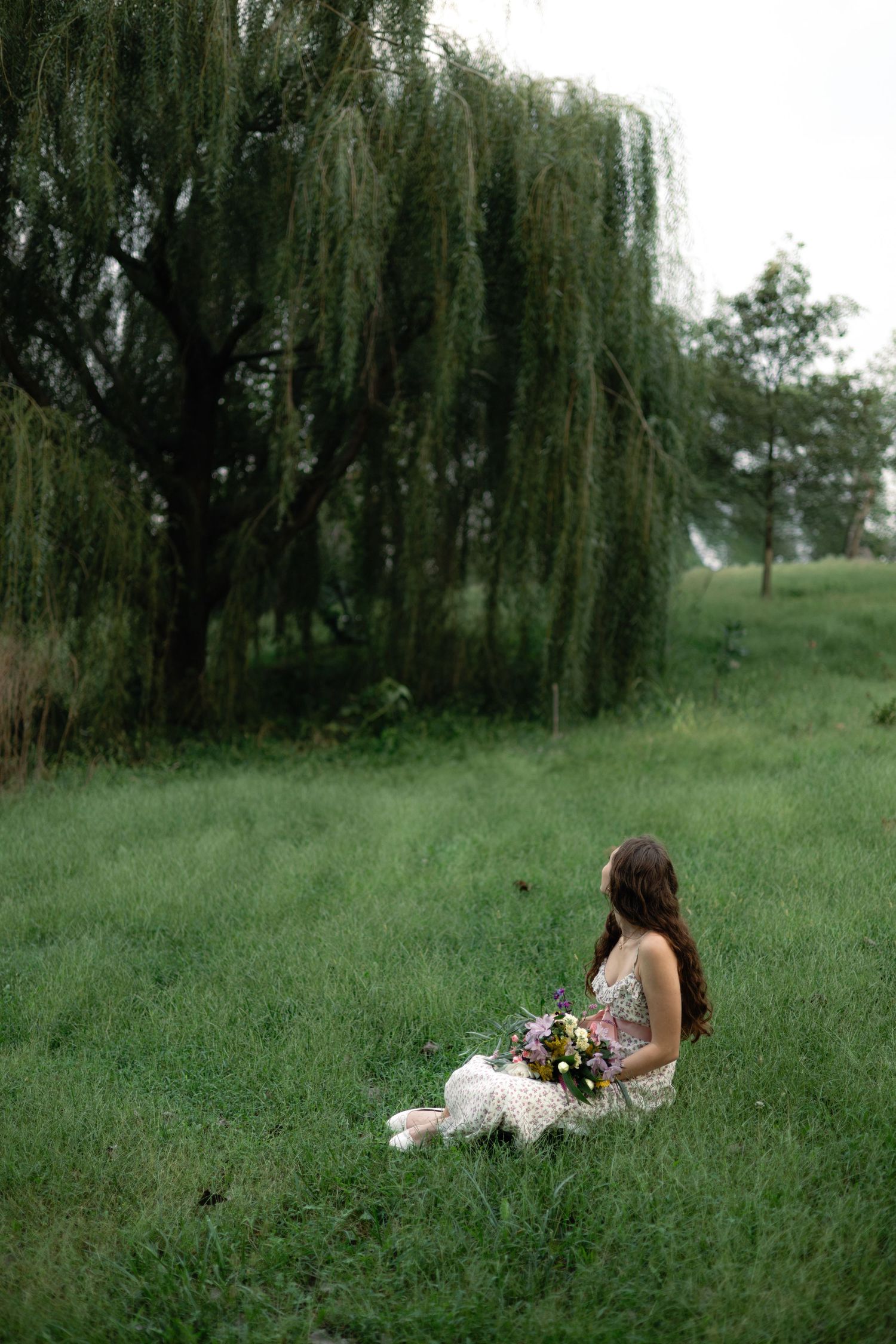 A young woman in a floral dress sits on lush green grass holding a bouquet near a weeping willow tree.