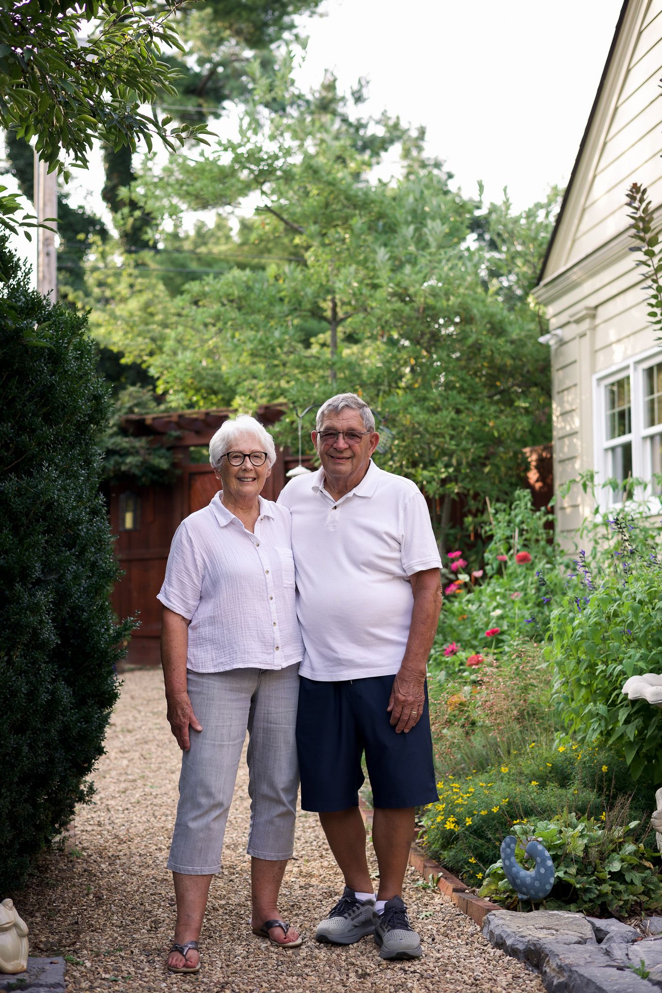 Senior couple smiling together in a lush backyard garden path beside their home on a sunny day.