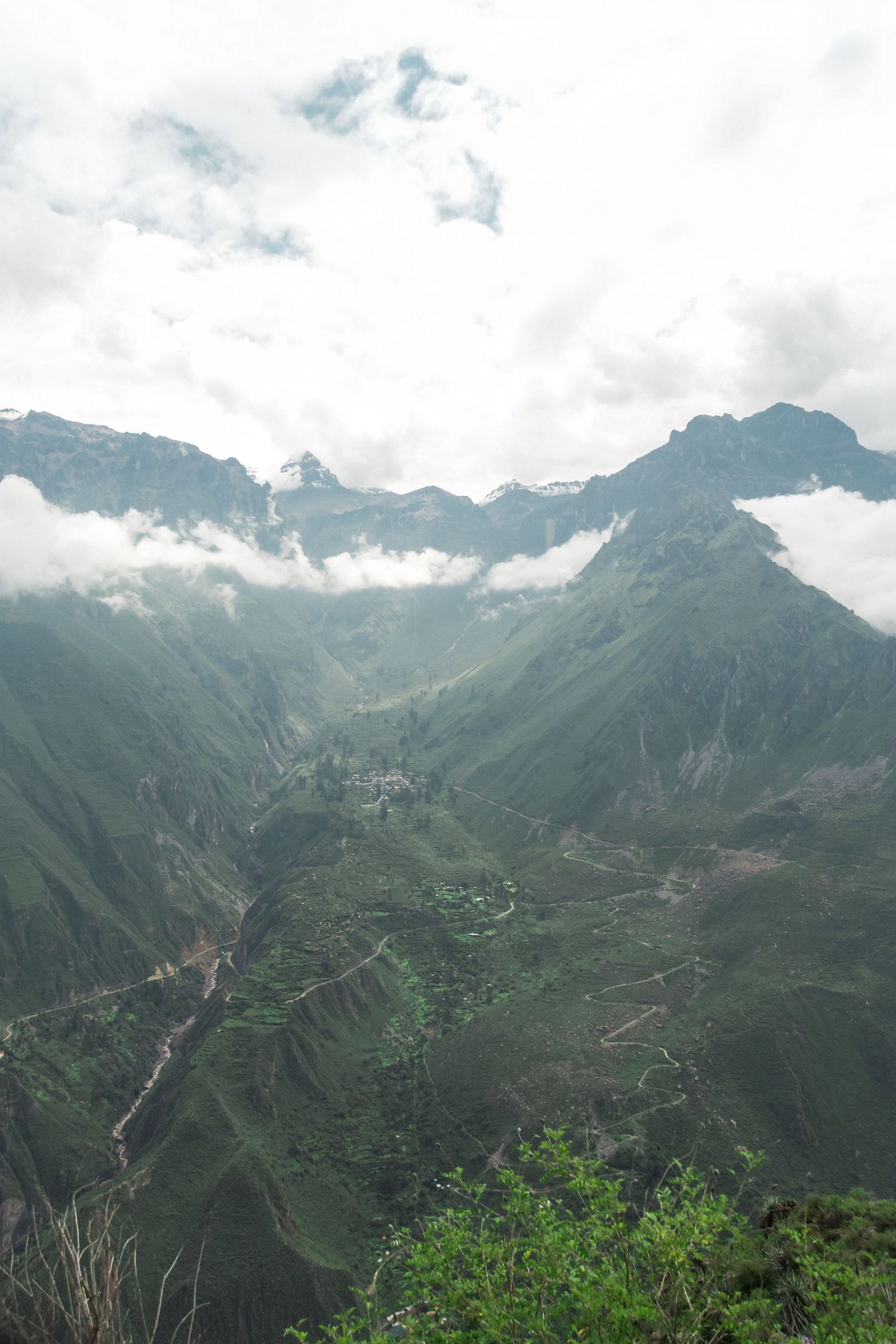 Aerial view of lush green mountain valley with terraced fields, winding river, and misty peaks under cloudy sky.