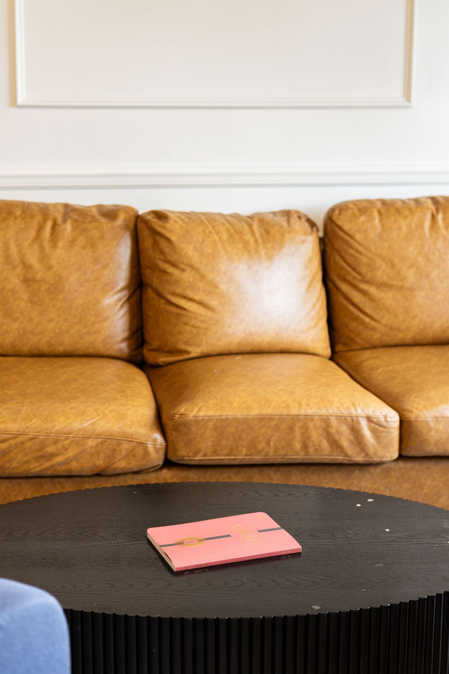 Tan leather sofa with three cushions beside a dark round coffee table holding a pink notebook.