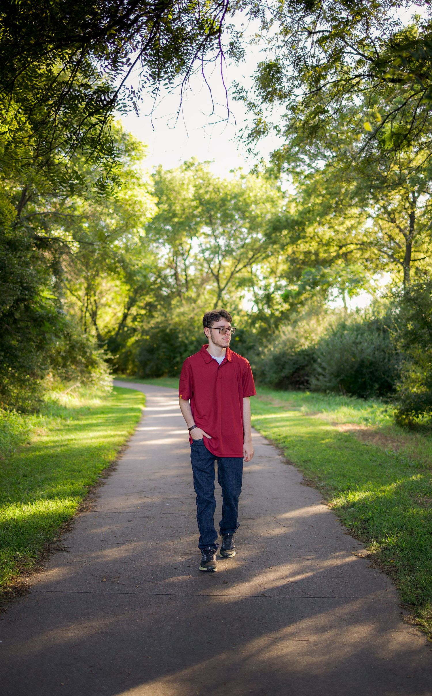 Young man in red polo shirt and jeans walking on a tree-lined park path on a sunny day.