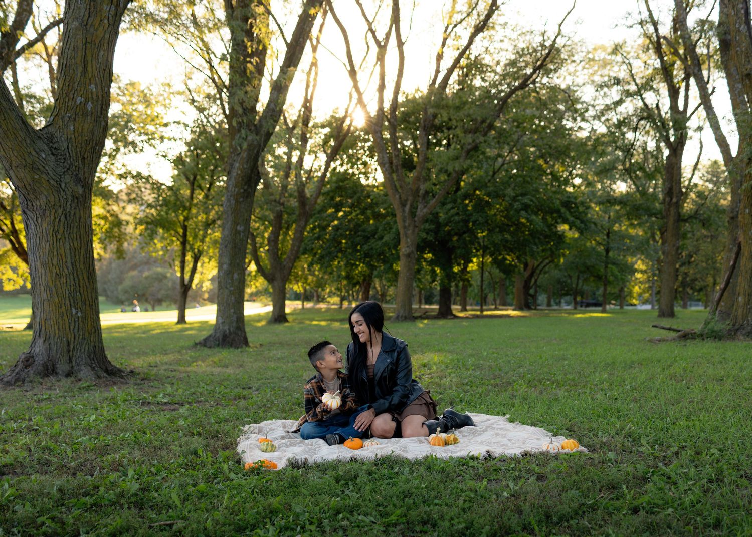 Mother and child sit on a white picnic blanket on a green lawn surrounded by tall trees in a sunlit park at golden hour.