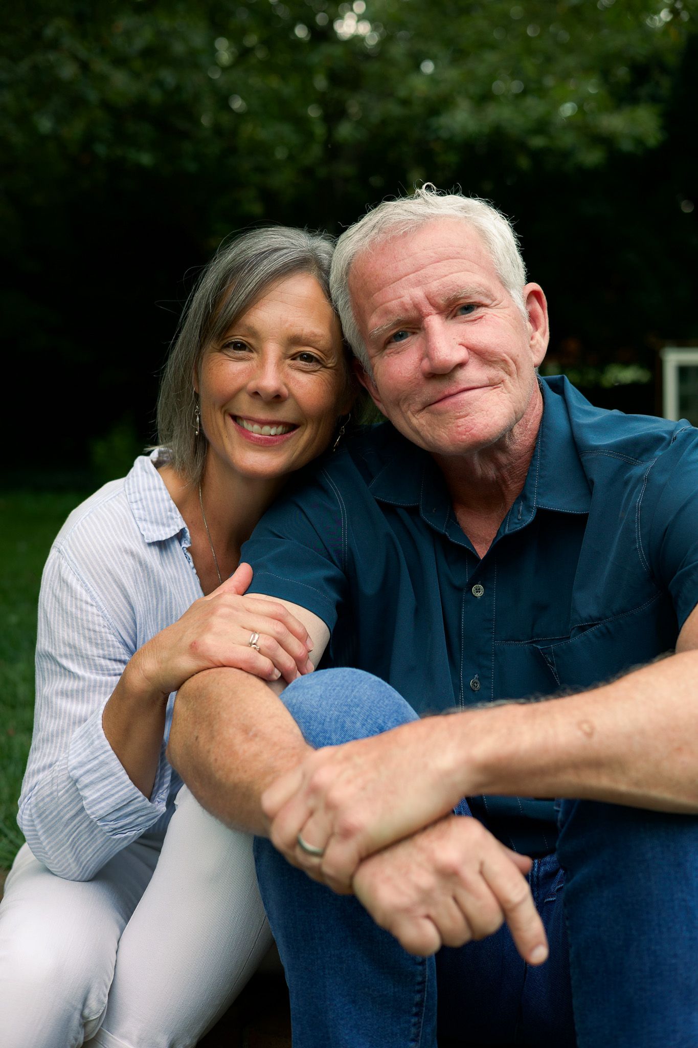 Happy senior couple sitting outdoors in a garden, woman embracing man from behind, both smiling warmly at the camera.