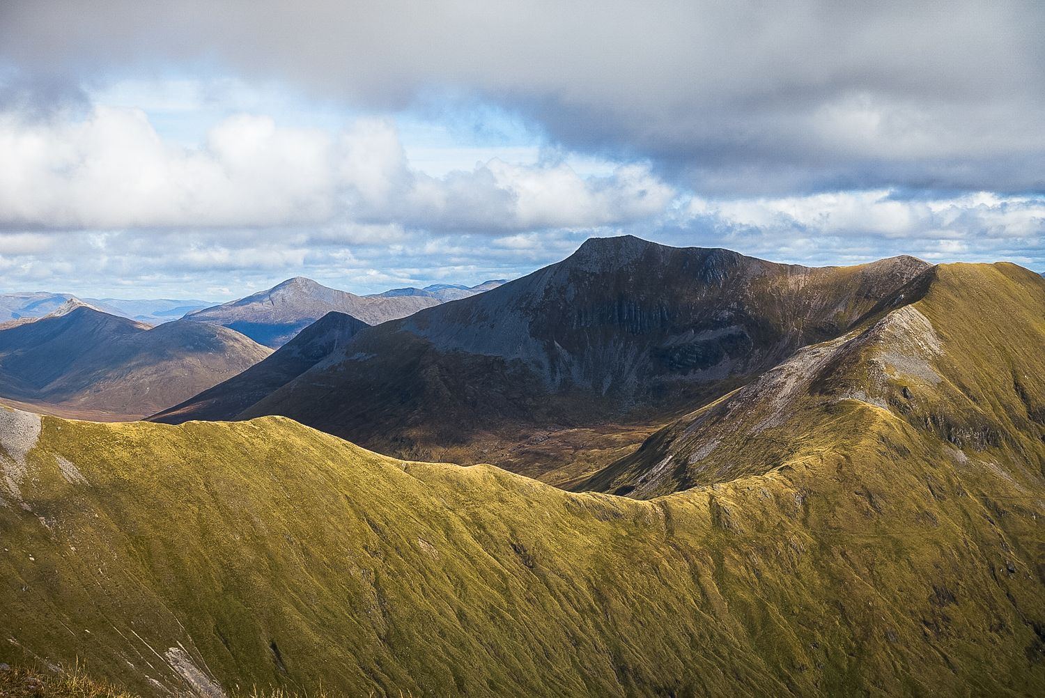Panoramic view of green rolling Scottish Highlands mountains under dramatic cloudy sky with sunlight illuminating the grassy slopes.