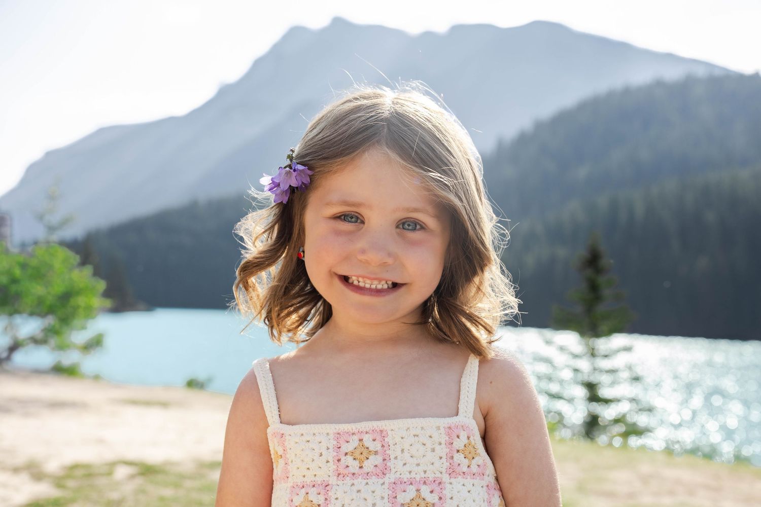 Smiling young girl with a purple flower hair clip stands near a mountain Lake Minnewanka on a sunny day in Banff