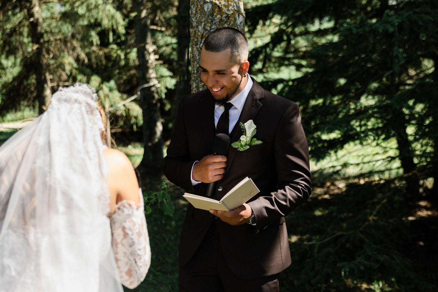 Groom in dark suit reads vows from a book to bride in lace veil during outdoor forest wedding ceremony.