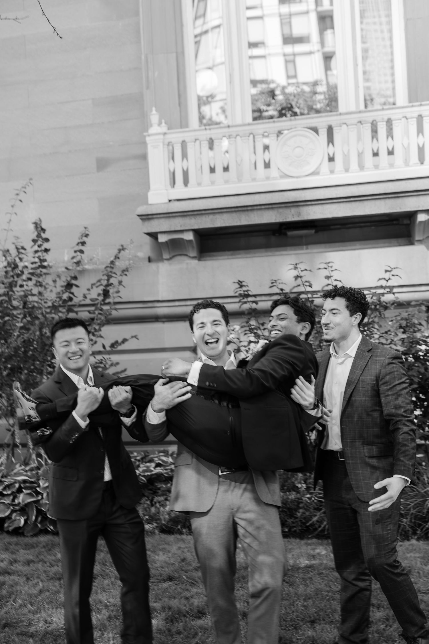 Groomsmen laughing and celebrating outdoors in suits near elegant building with balcony in black and white photo.