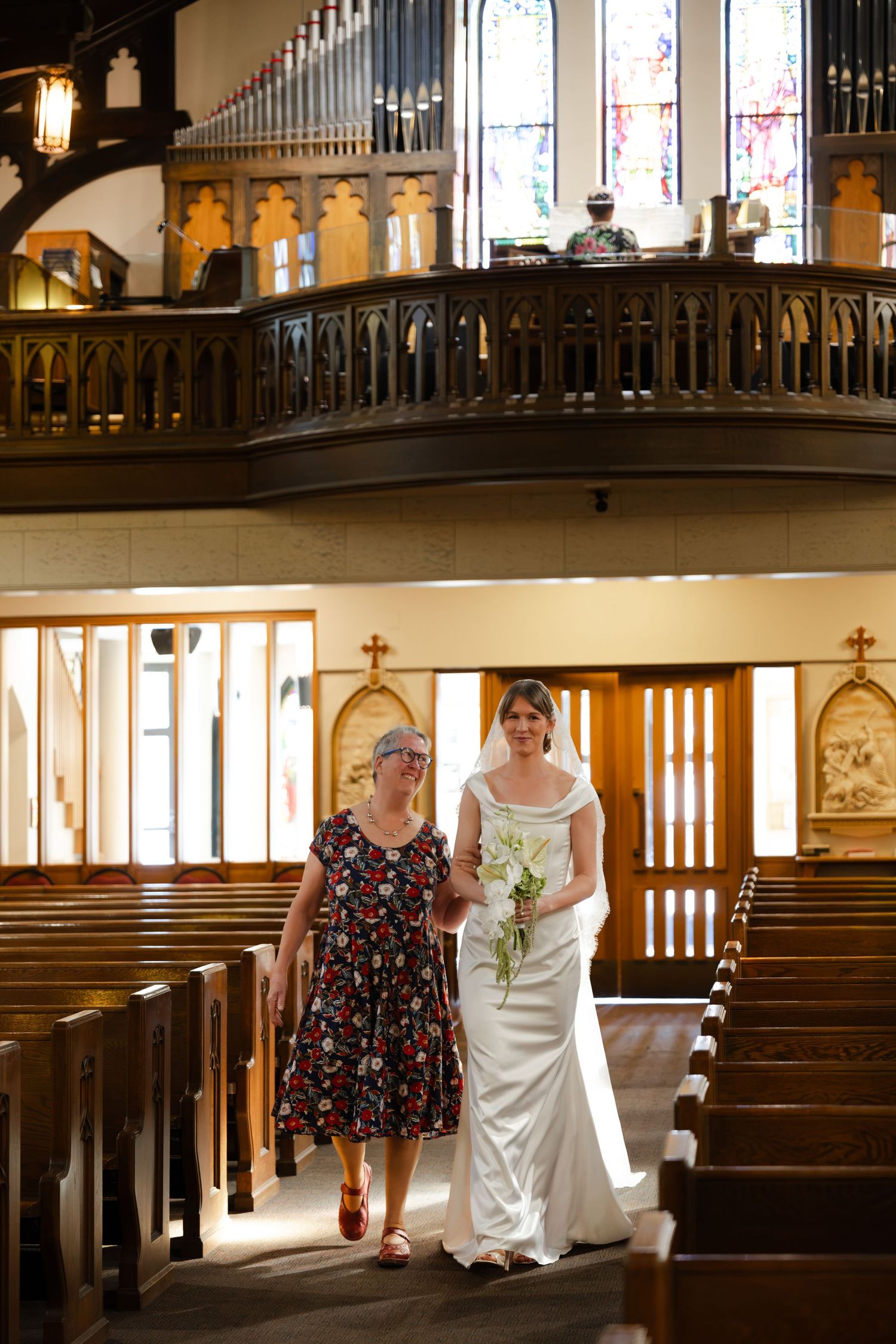 Bride in white gown walks down church aisle with older woman in floral dress, holding bouquet.