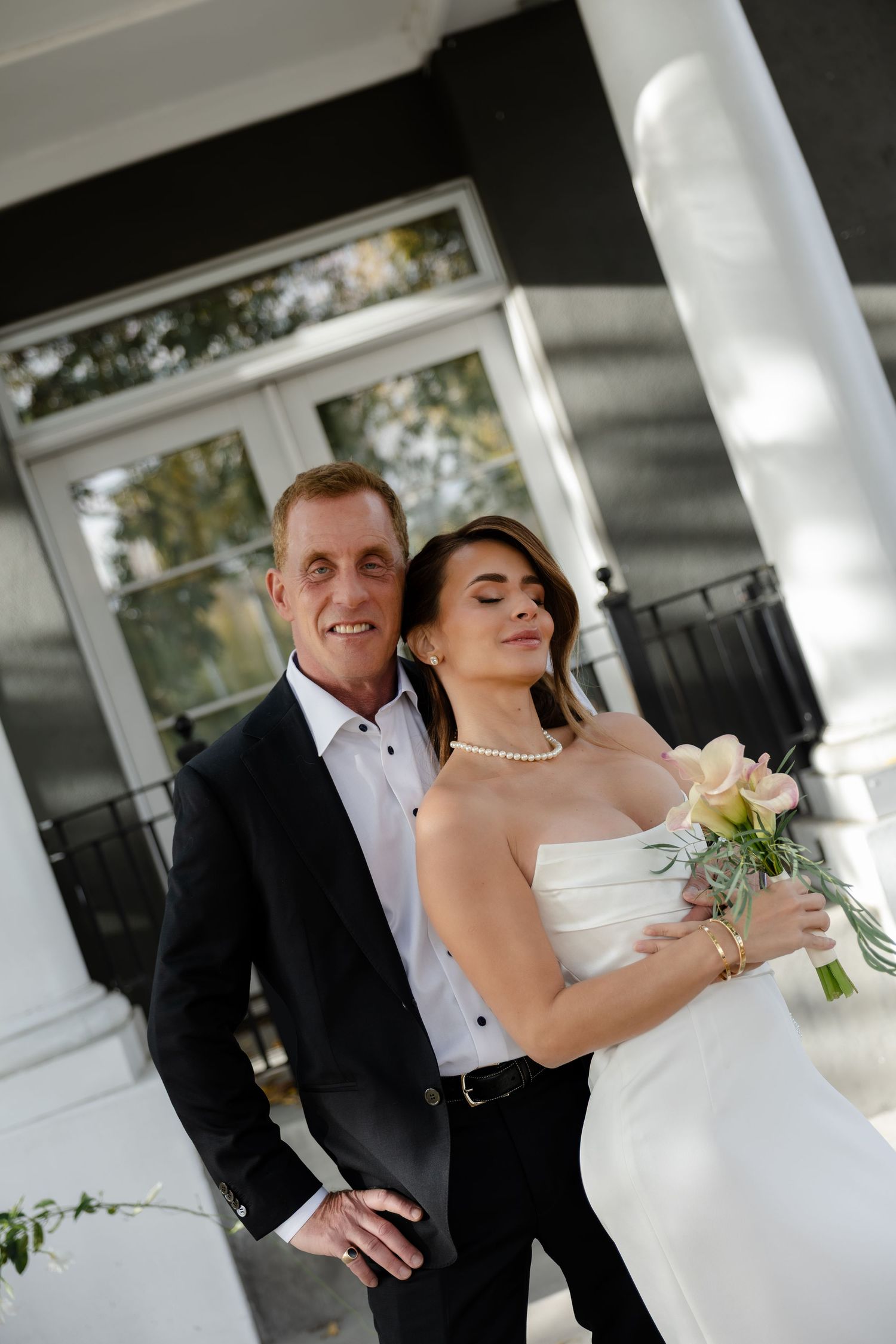 Bride in strapless white gown holding bouquet leans against groom in black suit on elegant porch.