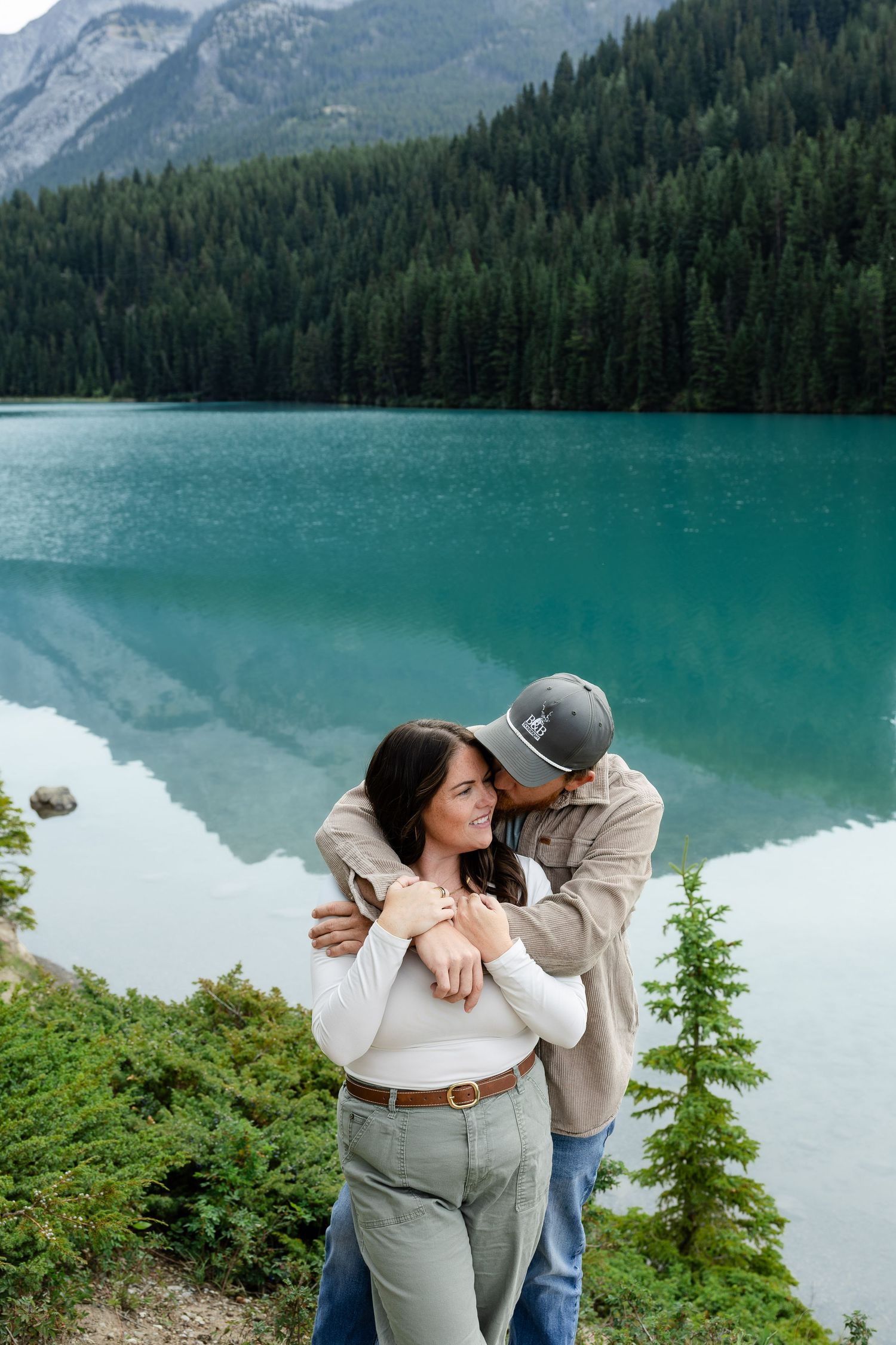 Couple embracing by a turquoise mountain lake with evergreen forest backdrop.