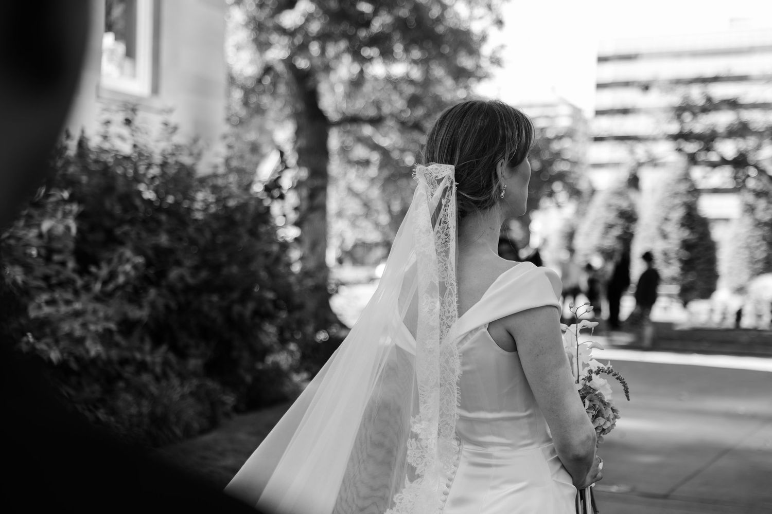 Black and white photo of a bride in a white gown and veil, seen from behind, holding a bouquet outdoors.
