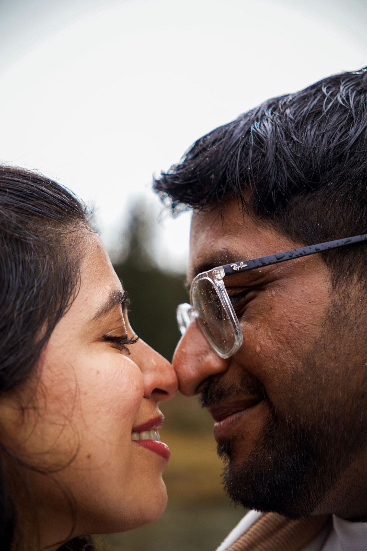 Close-up portrait of a smiling couple touching noses, man wearing glasses, sharing a romantic moment outdoors.