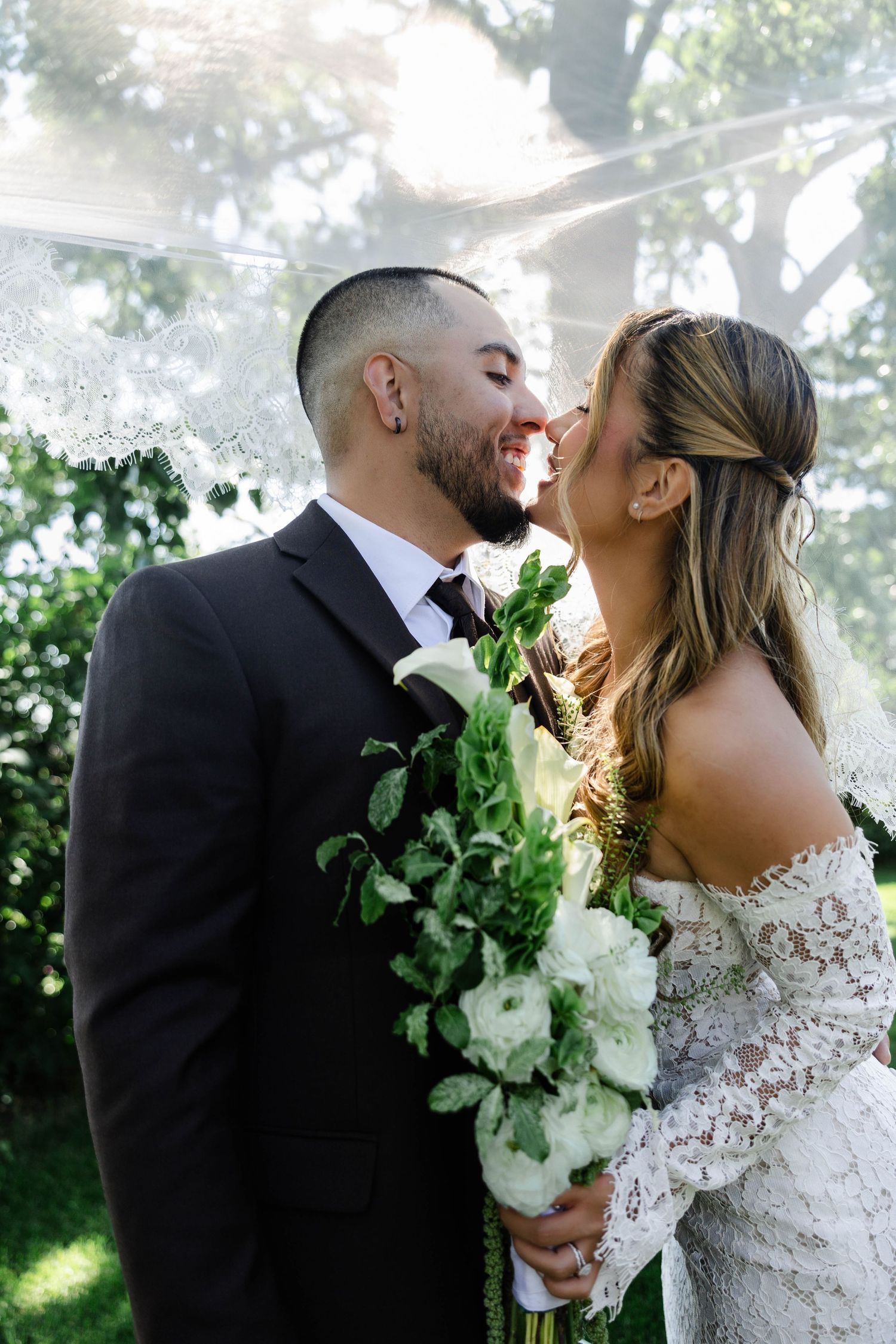 Bride and groom share a romantic moment under a veil, bride holding white floral bouquet outdoors.