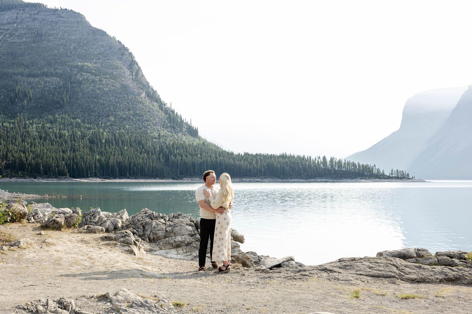 Couple embracing by a serene mountain lake with rocky shores and forested peaks in the background.Romantic couple in Minnewanka Lake in Banf