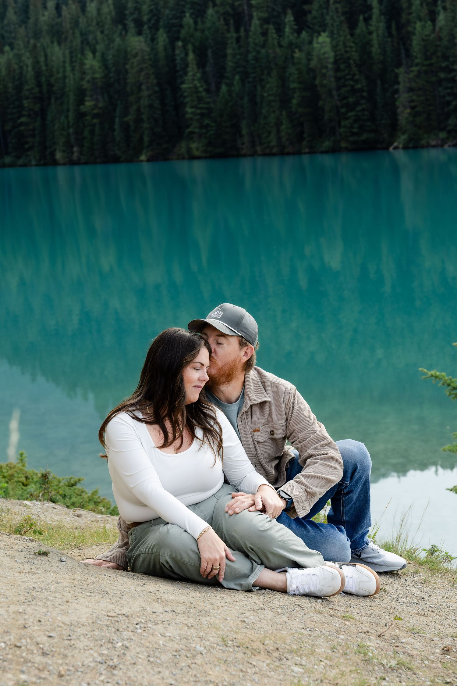 Couple sitting by a turquoise mountain lake, man kissing woman's head, surrounded by pine trees.