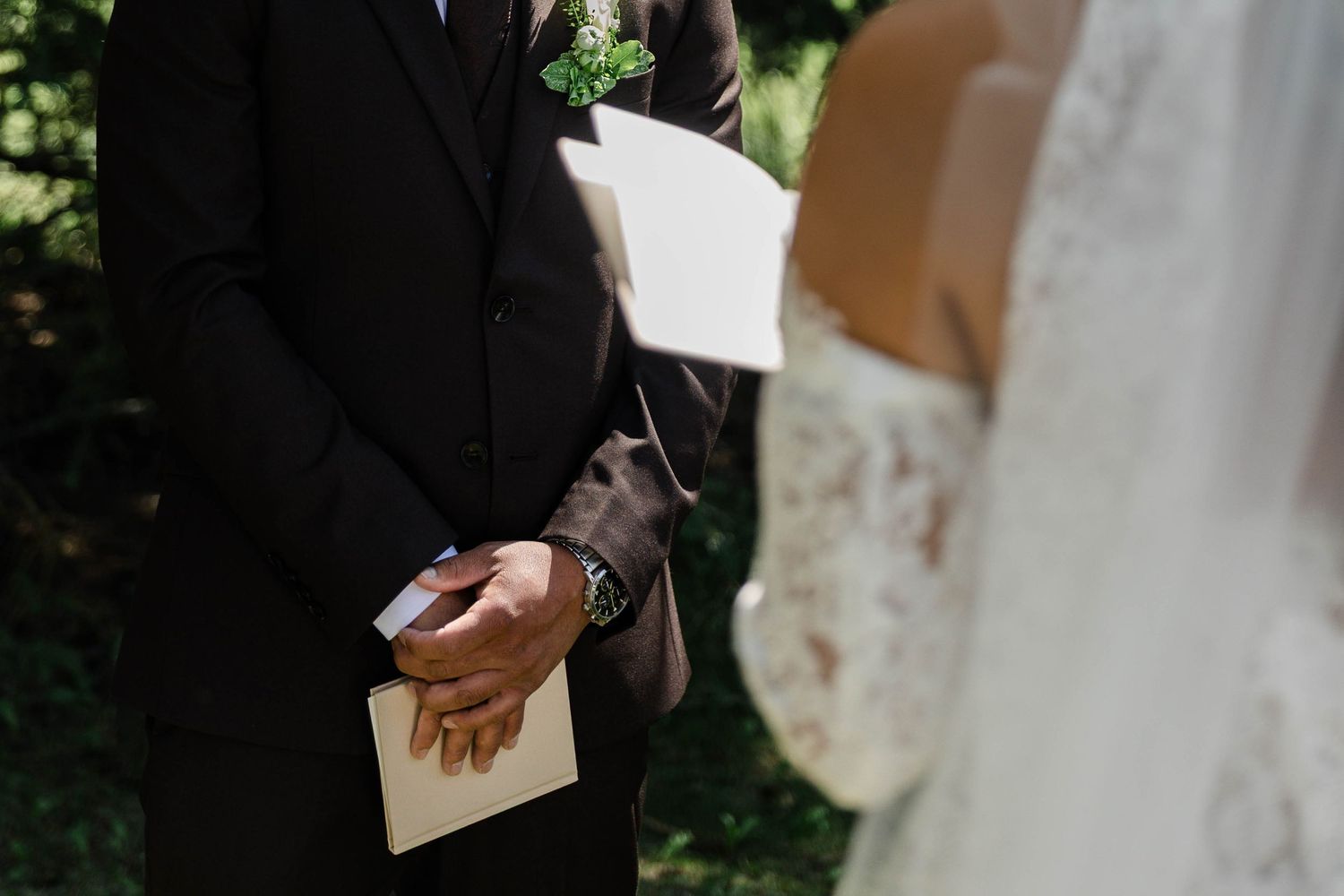 Groom in black suit holding vows card during outdoor wedding ceremony with bride in lace dress.