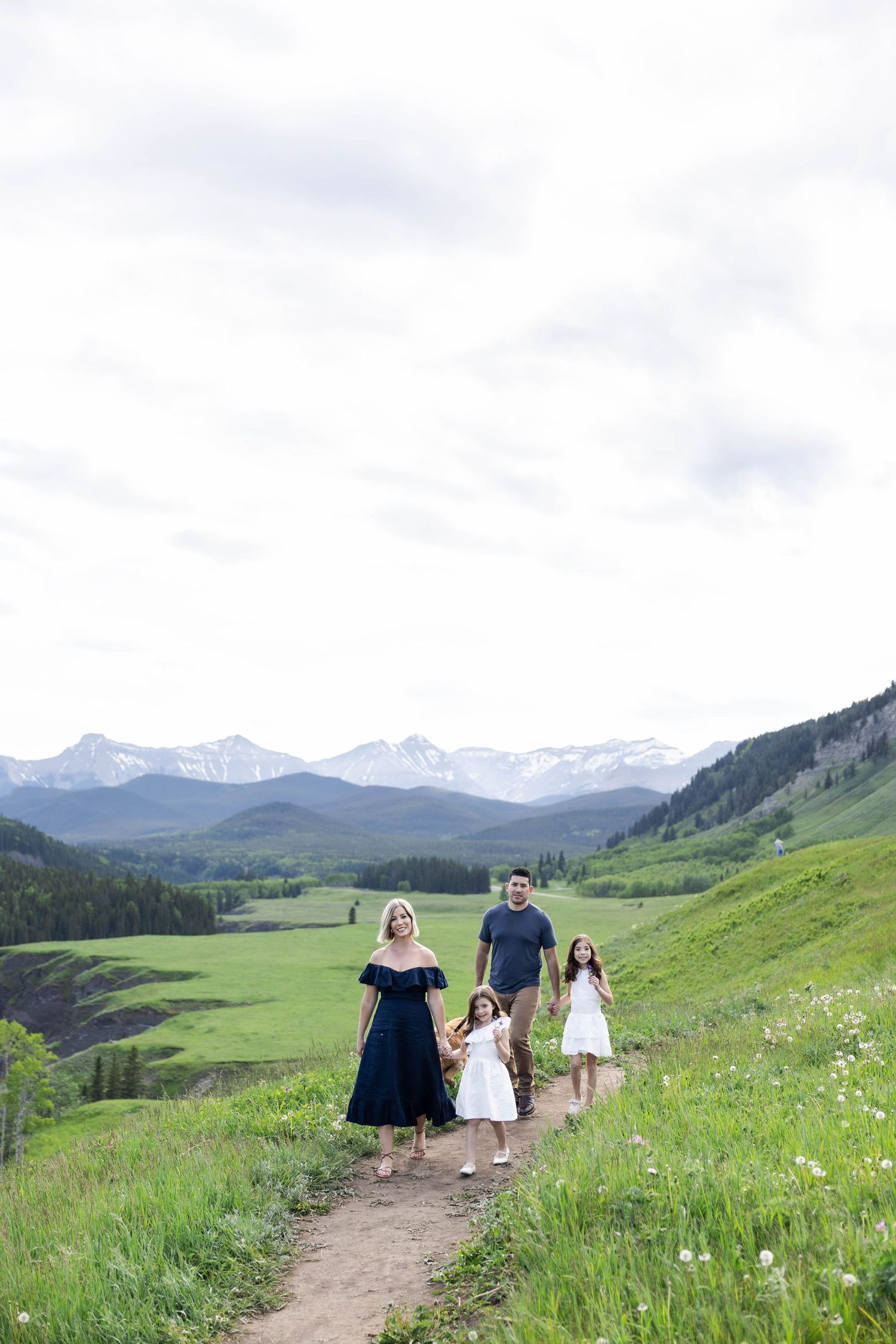 Family of four walking on a mountain trail with snow-capped peaks and green meadows in the background.