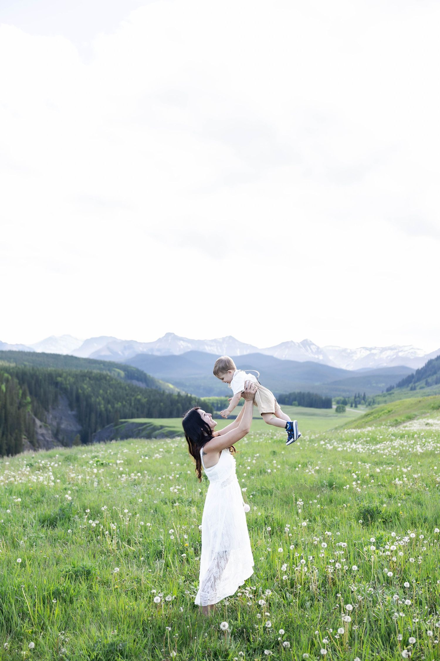 A mother in a white dress lifts her baby in a sunny mountain meadow with wildflowers and snow-capped peaks.