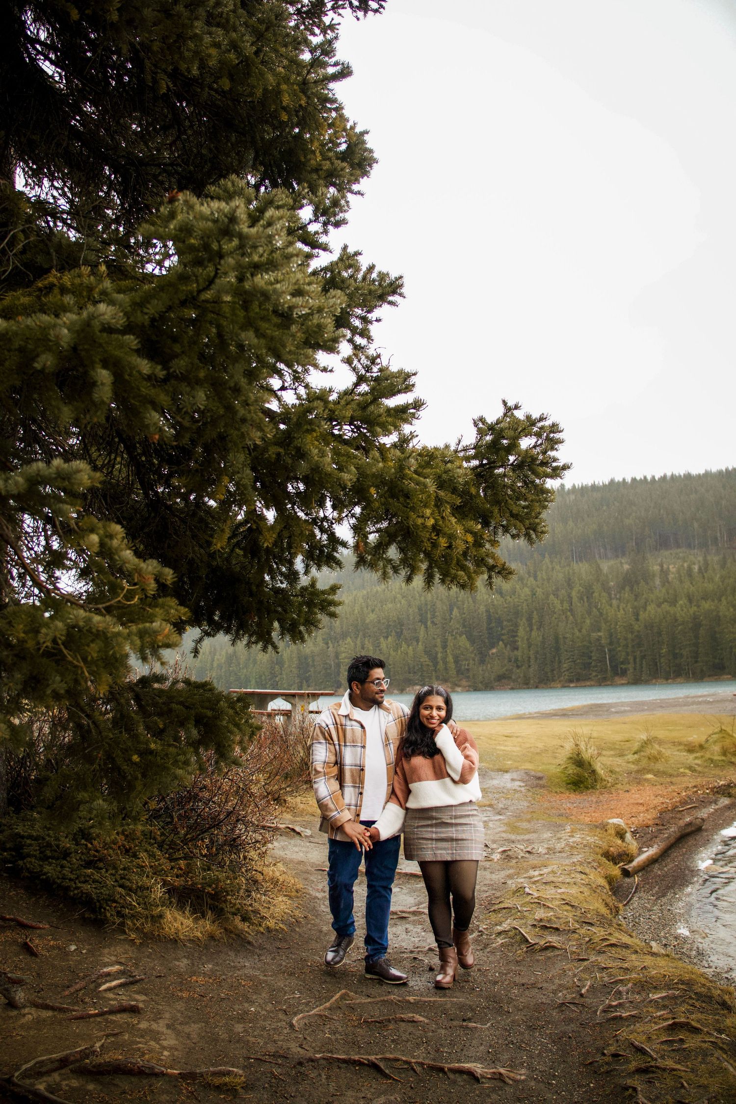 Couple holding hands walking on a forest path beside a mountain lake surrounded by tall evergreen trees.