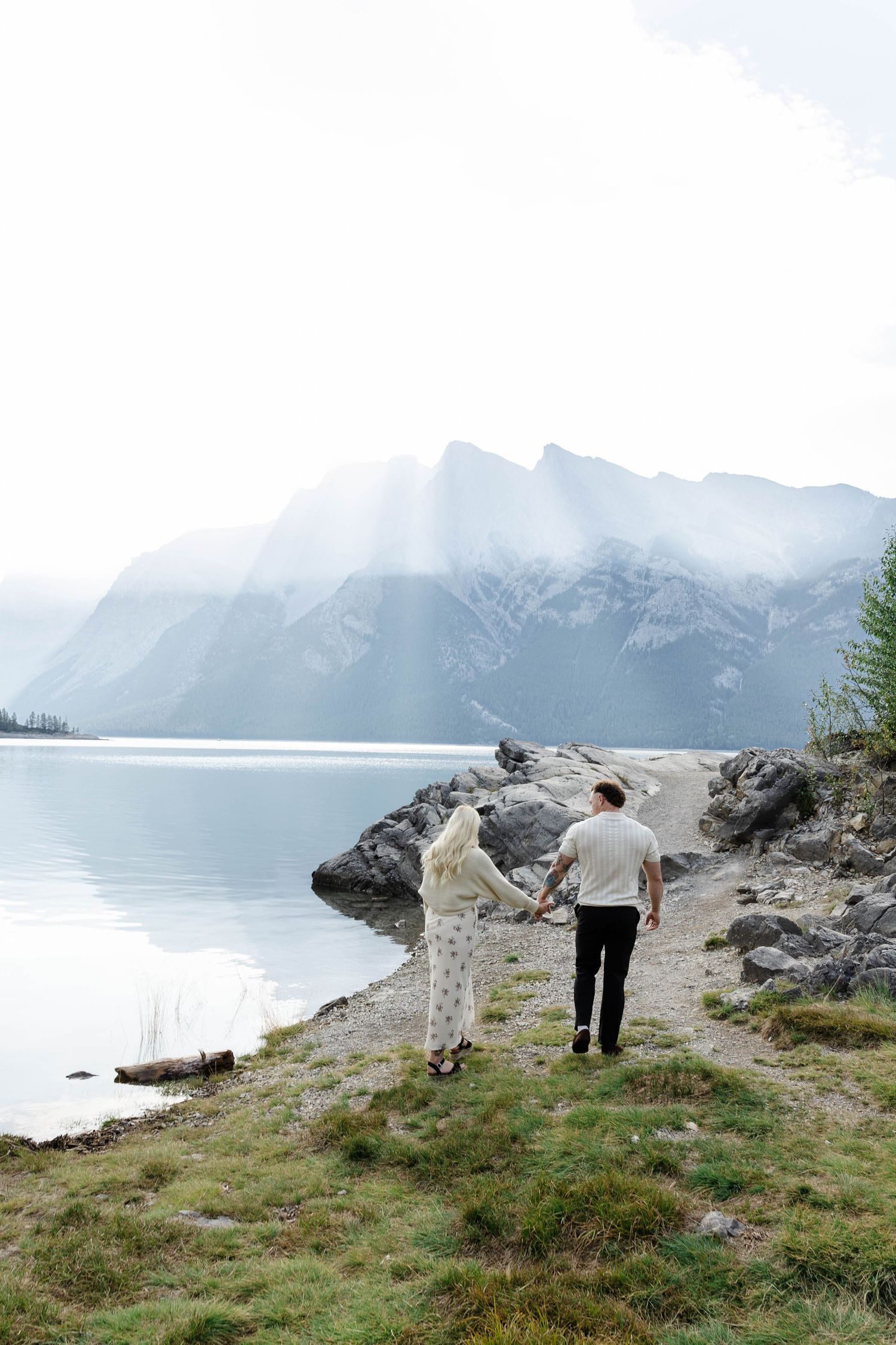 Couple holding hands walking toward a misty mountain lake surrounded by rocky shores and evergreen trees.Romantic couple in Minnewanka Lake