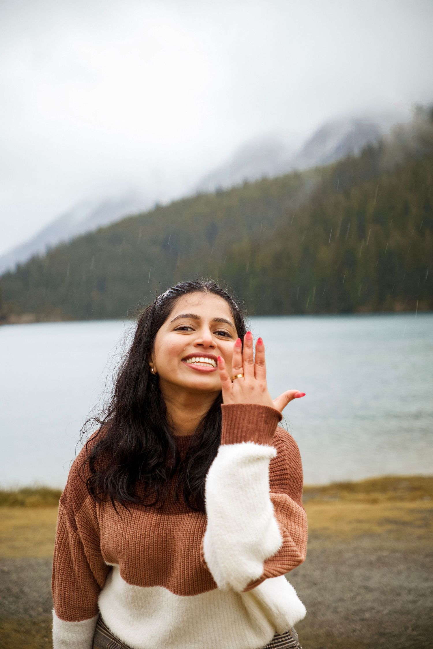 Smiling young woman in brown sweater posing near a misty mountain lake, hand raised to face.