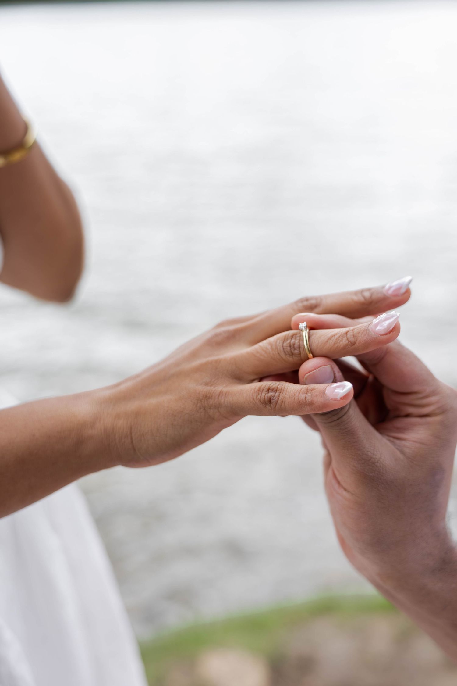Close-up of a groom placing a gold wedding ring on the bride's finger near a serene waterfront.
