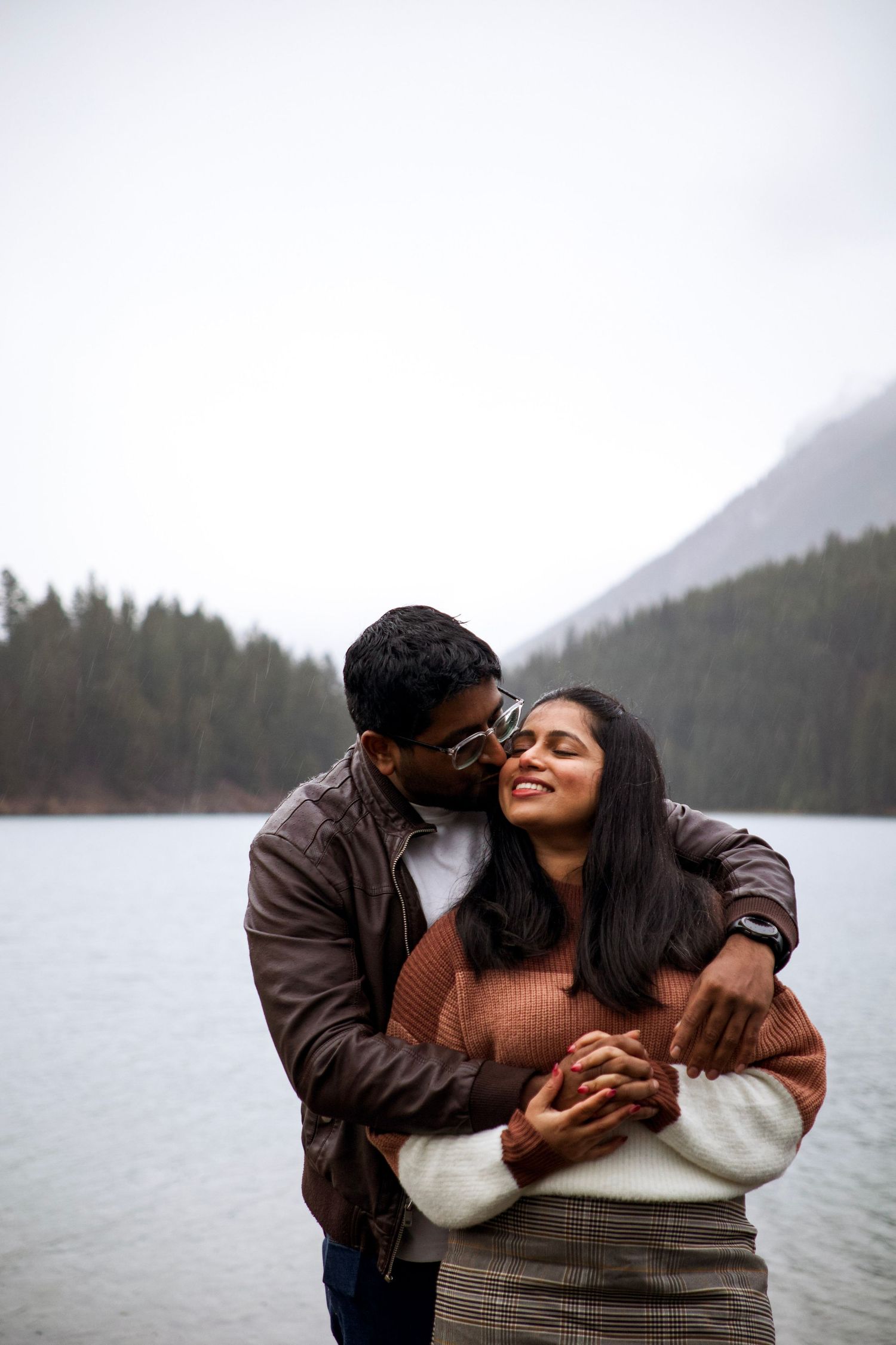 Couple embracing by a misty mountain lake, man kissing woman's cheek as she smiles with eyes closed in Banff