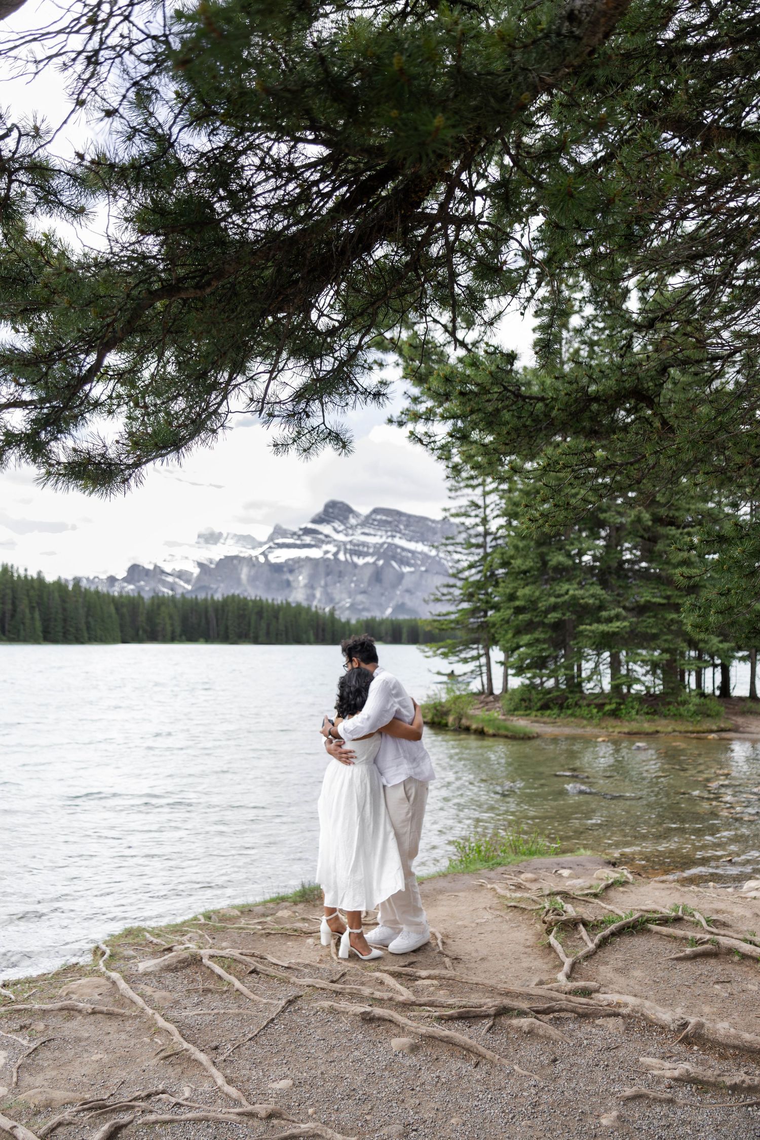 Couple embracing in wedding attire by a mountain lake surrounded by pine trees in Banff National Park.