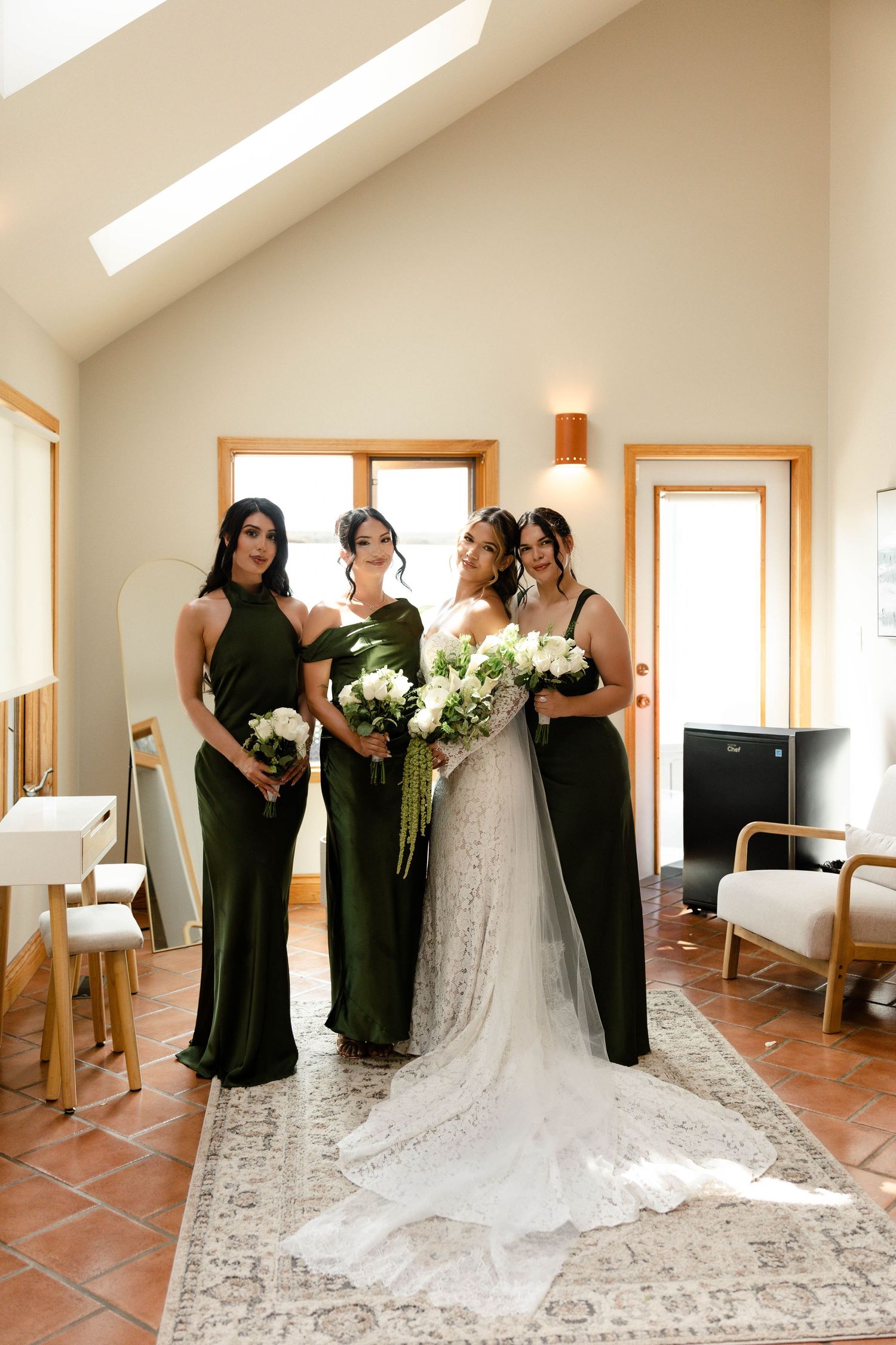 Bride in lace gown with two bridesmaids in olive green dresses holding white bouquets indoors.
