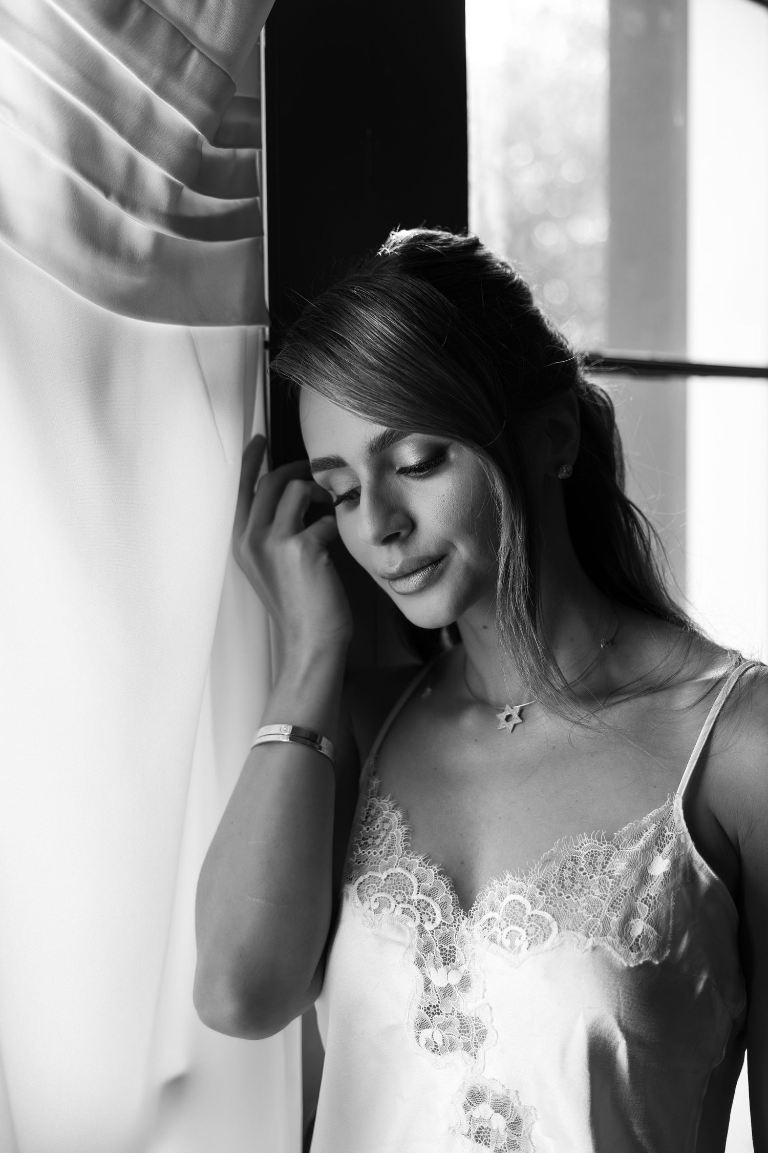 Black and white portrait of a woman in lace camisole leaning against wall by window, eyes closed, smiling softly.