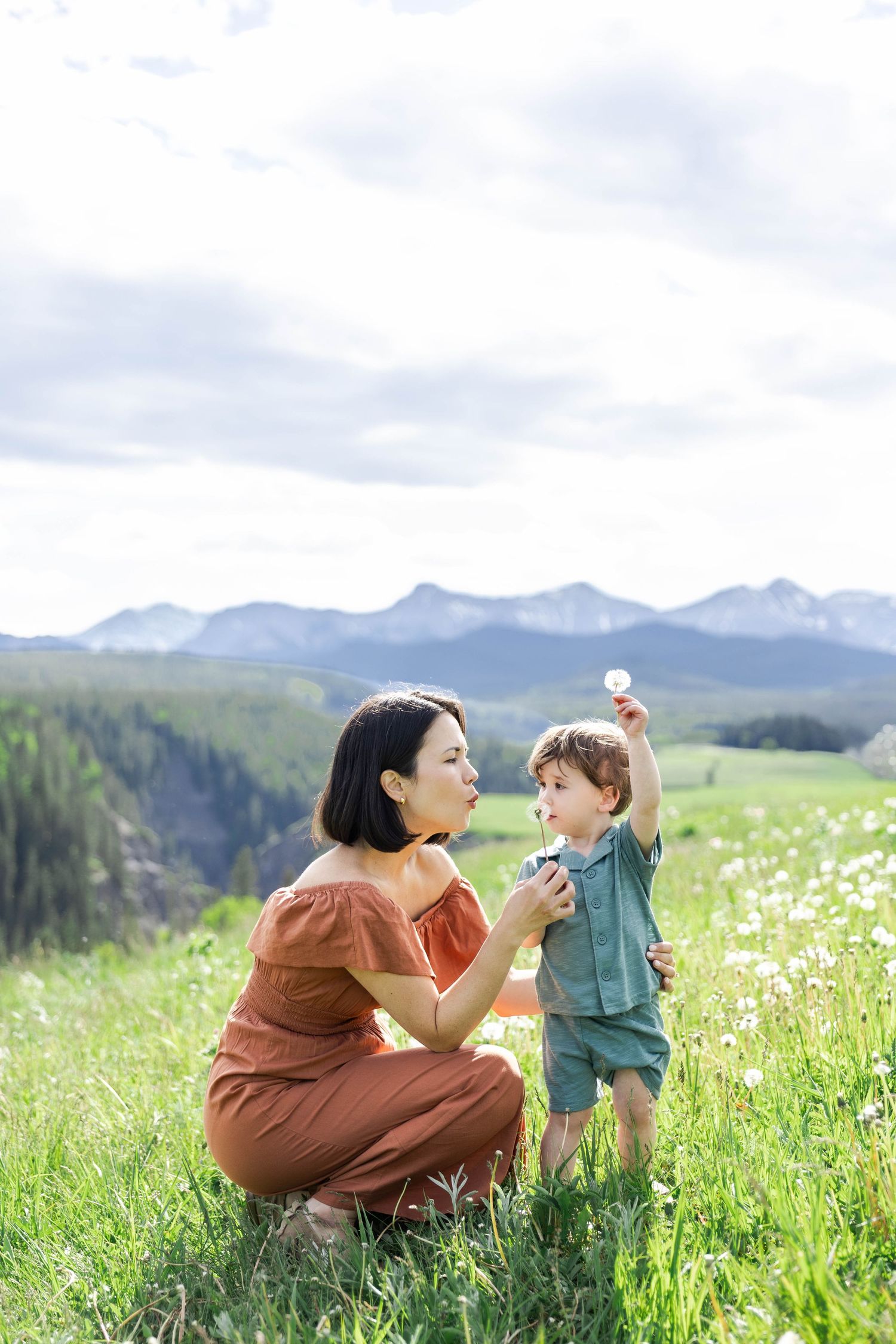 Mother and toddler blowing dandelions in a green meadow with mountain views on a cloudy day.