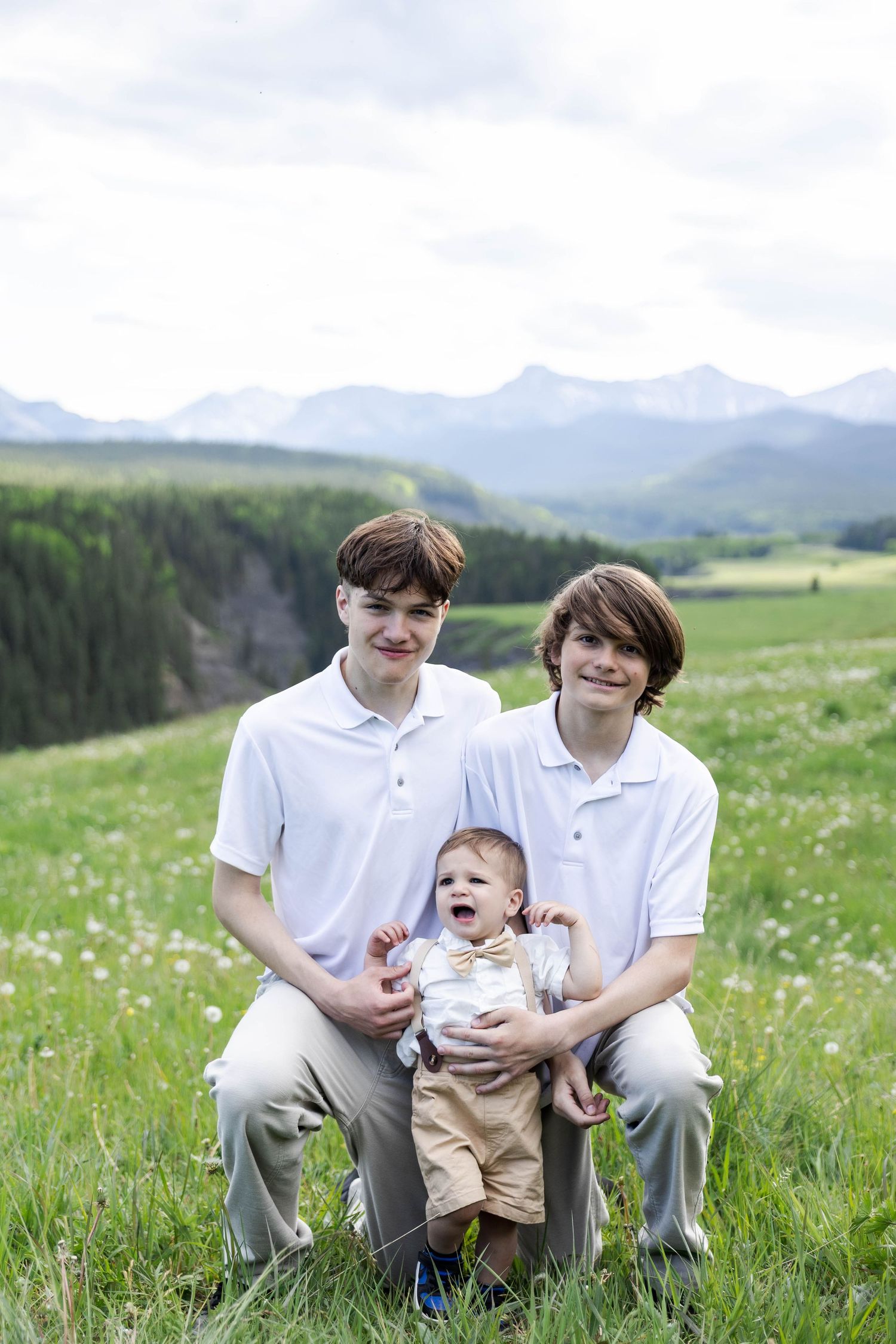 Two teenage boys in white polo shirts hold a laughing baby in a green mountain meadow.