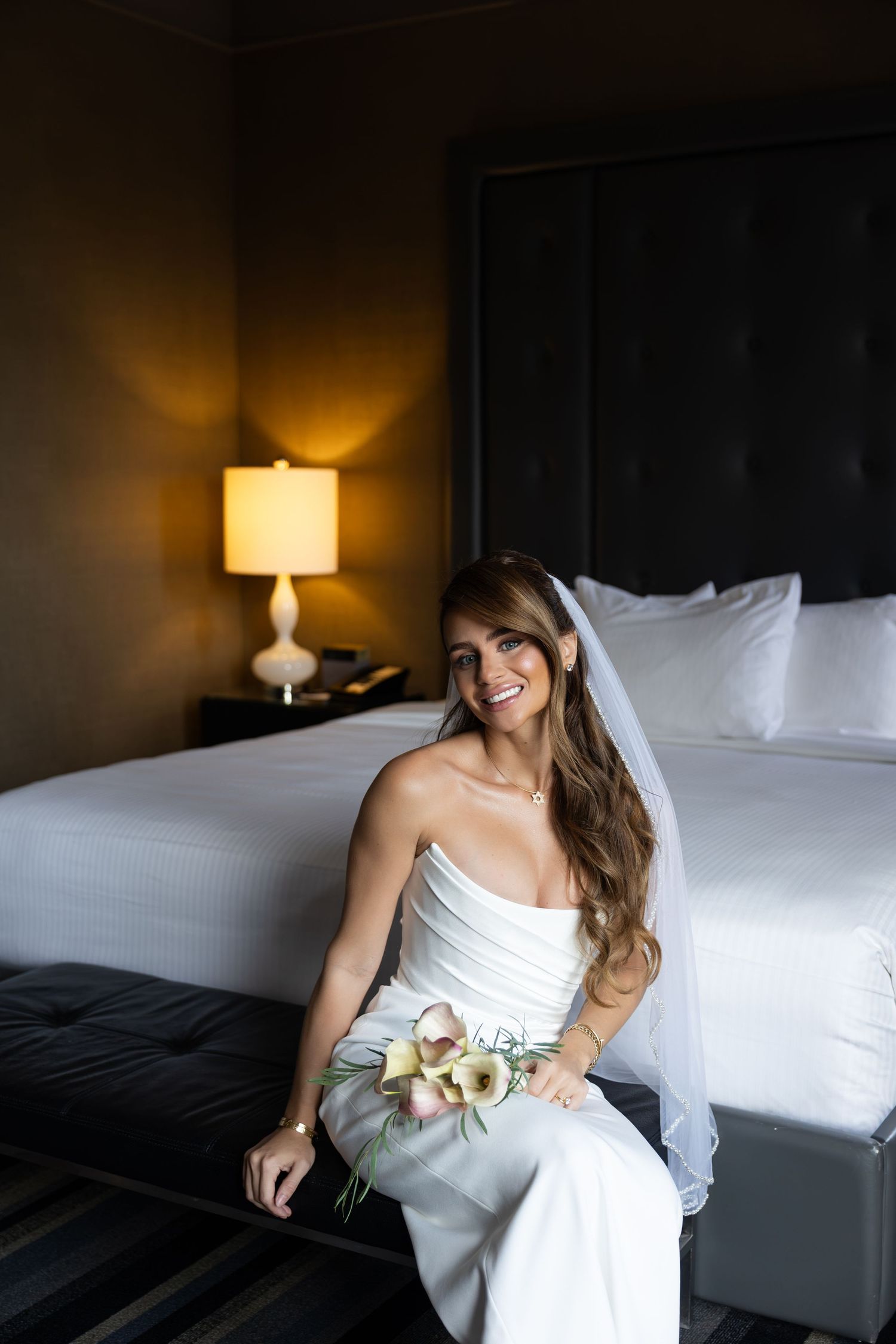 Smiling bride in white strapless gown and veil sits on hotel bed bench holding delicate floral bouquet.