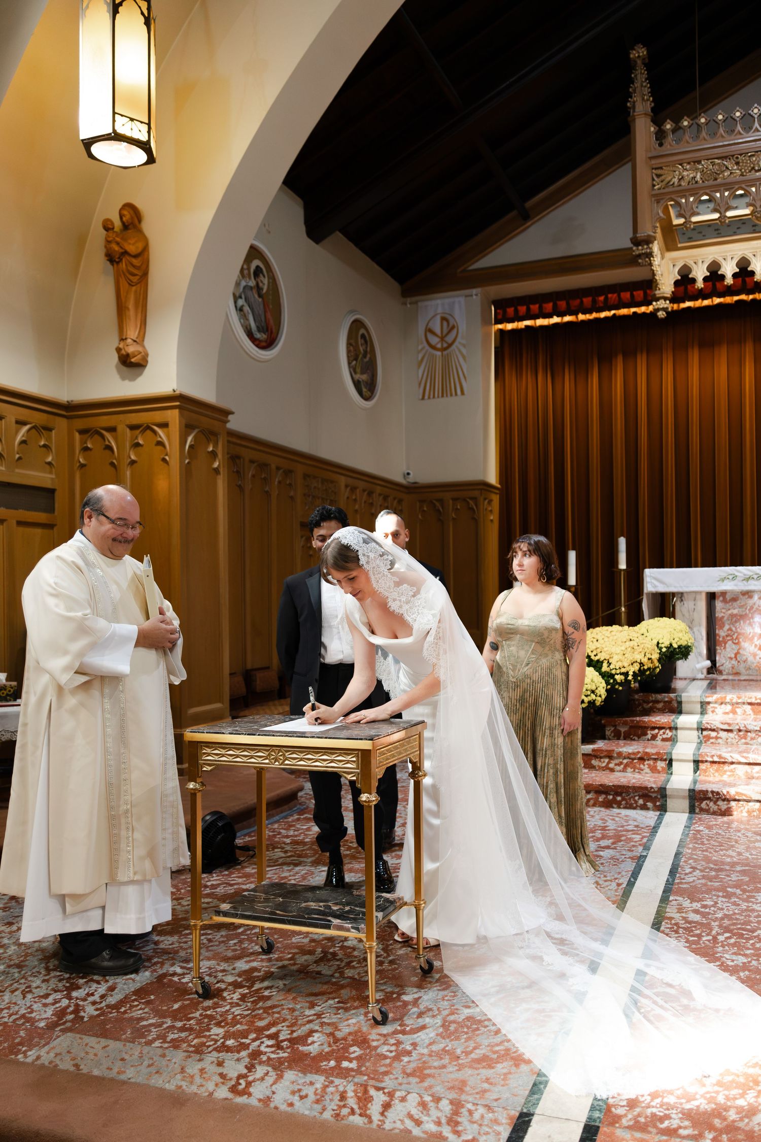 Bride signing wedding registry at church altar with priest and guests during Catholic wedding ceremony.
